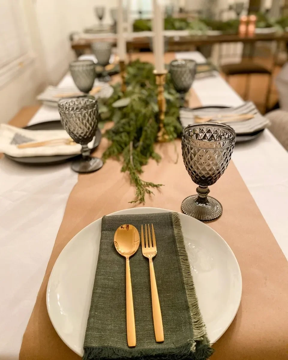 Wedding table styling - Elegant dining table set with black goblets, gold flatware on a green cloth napkin, and a white plate, with a green garland centrepiece and a gold candlestick.