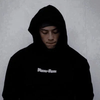 A person wearing a black hoodie and a black cap, looking downward, against a plain light background.