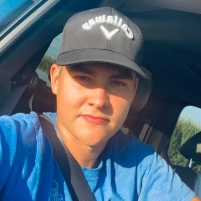Young man wearing a black Callaway cap and blue shirt, sitting in a vehicle with sunlight on his face.