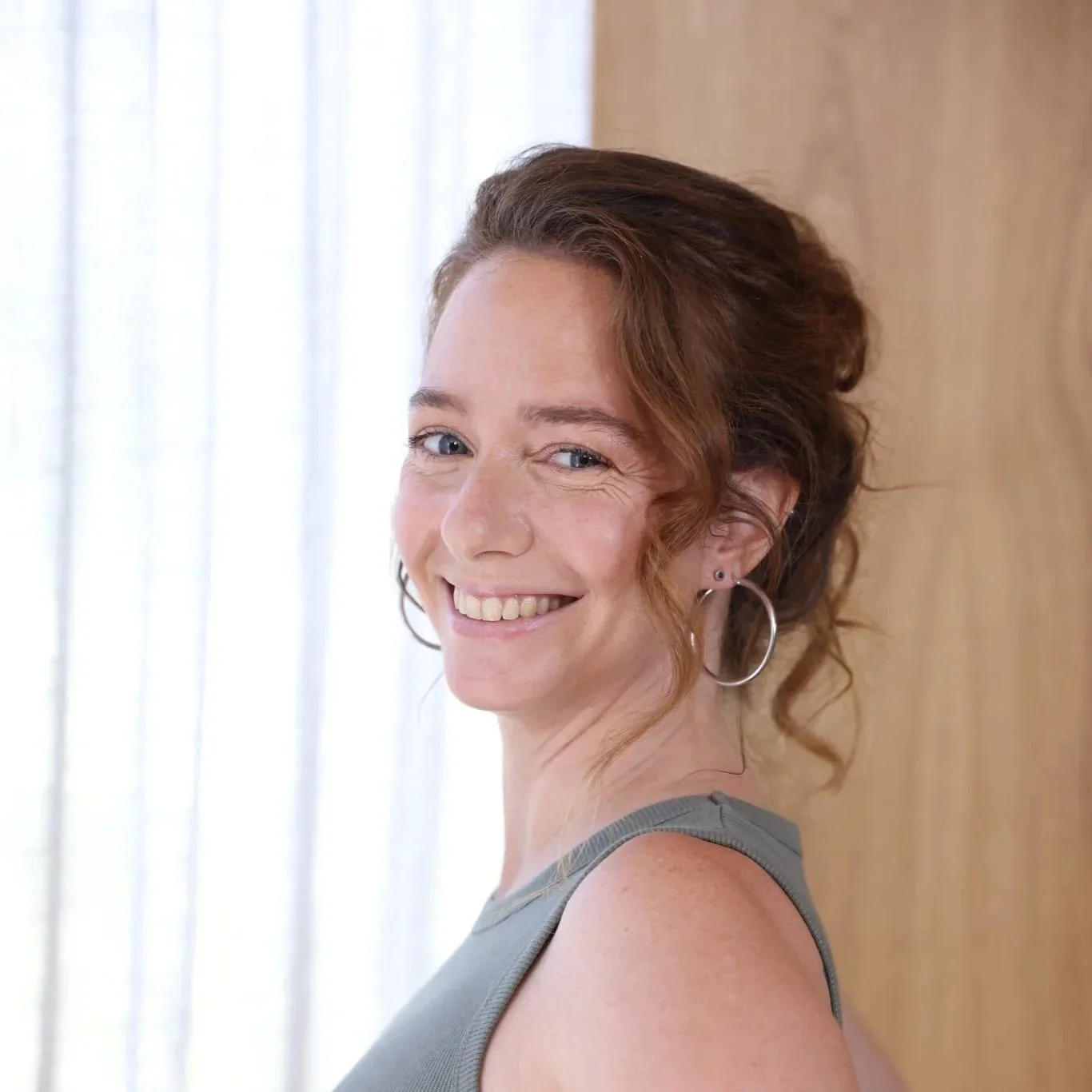Portrait of Rena looking at the camera with her hair in an updo wearing a green tank top and big, round silver earrings with a white curtain in the background and a brown wall