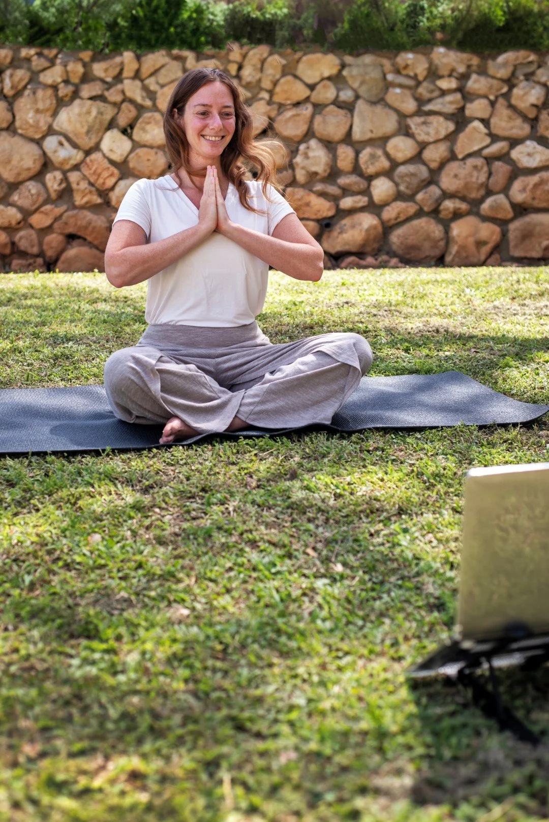 Photo of Rena with her hands in the Namaste position together in front of her heart, she is seated on a Yoga mass which is on the grass with a rock wall in the background, Rena is smiling