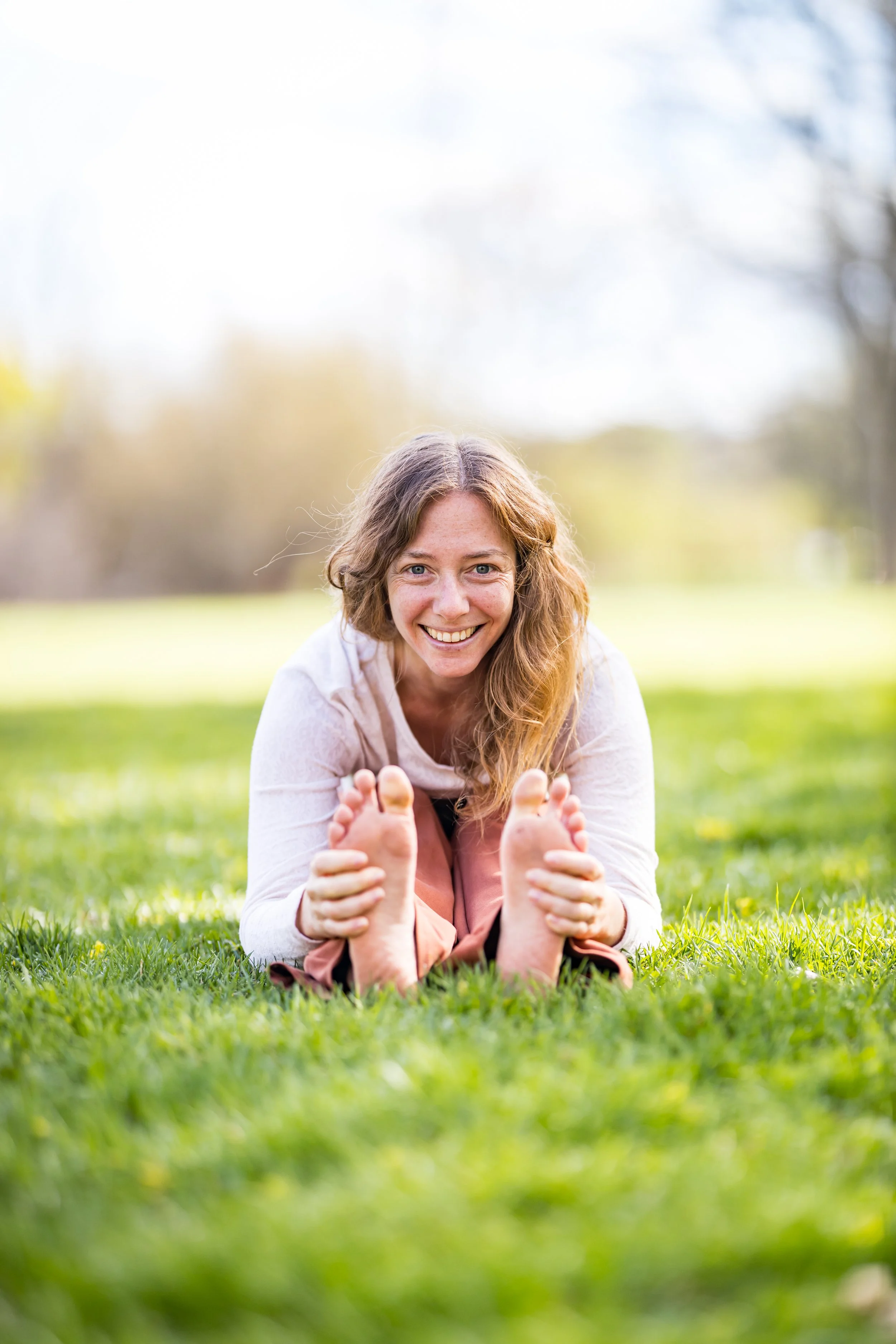 Photo of Rena sitting in the grass with her hands on. herfeet in a Yoga pose while she smiles and stares at the camera
