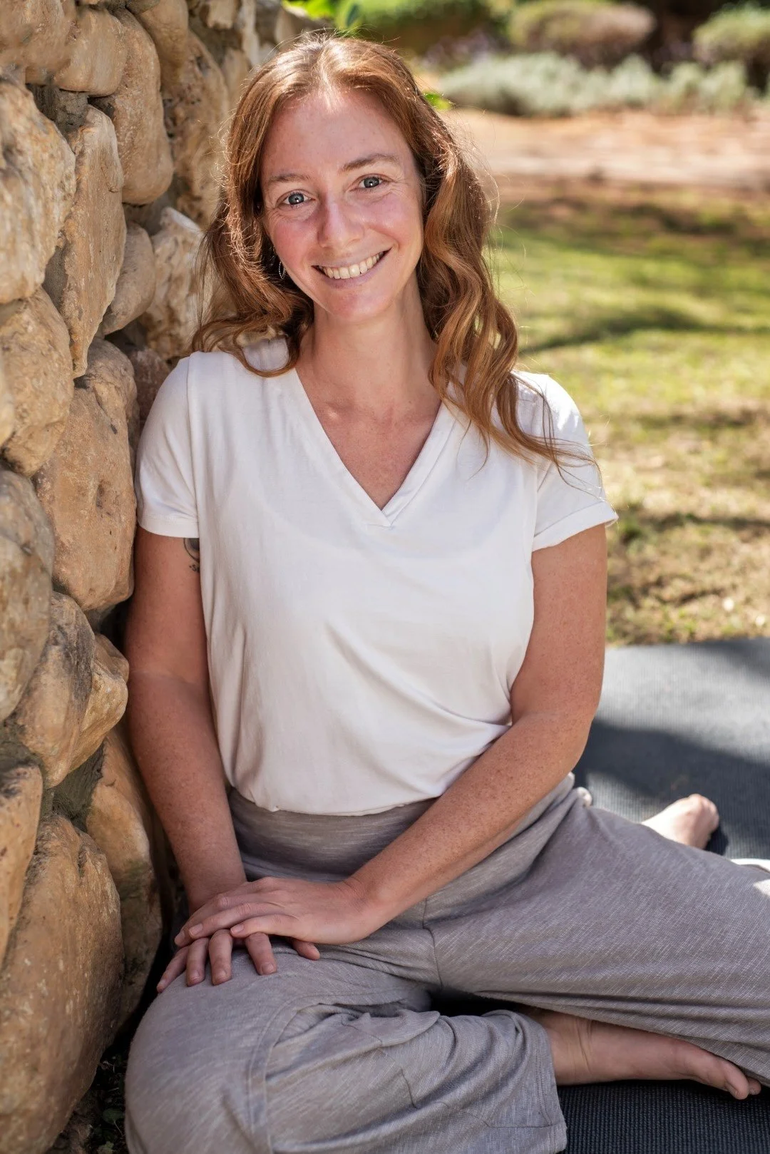 Photo of Rena in a white t-shirt leaning against a rock wall smiling and staring directly at the camera
