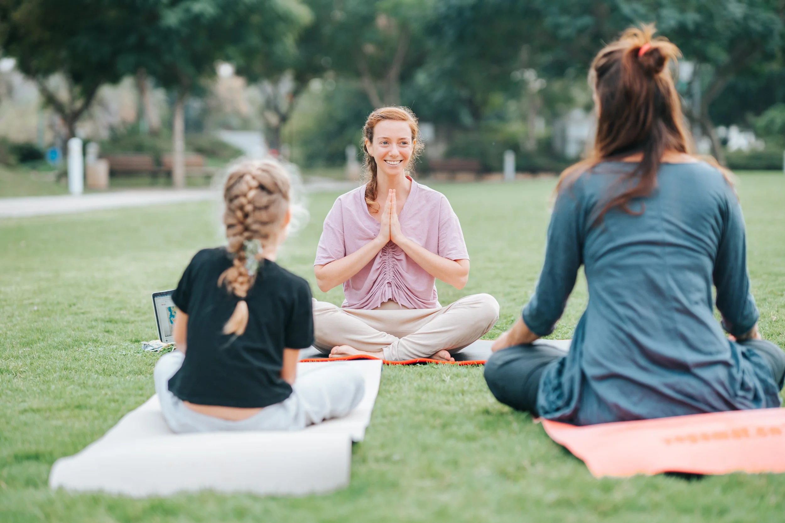 Picture of Rena teaching a Yoga class in the park to a young girl and a woman who are sitting on Yoga mats with their backs to the camera. Grass and trees surround them and Rena is smiling as she looks at her students