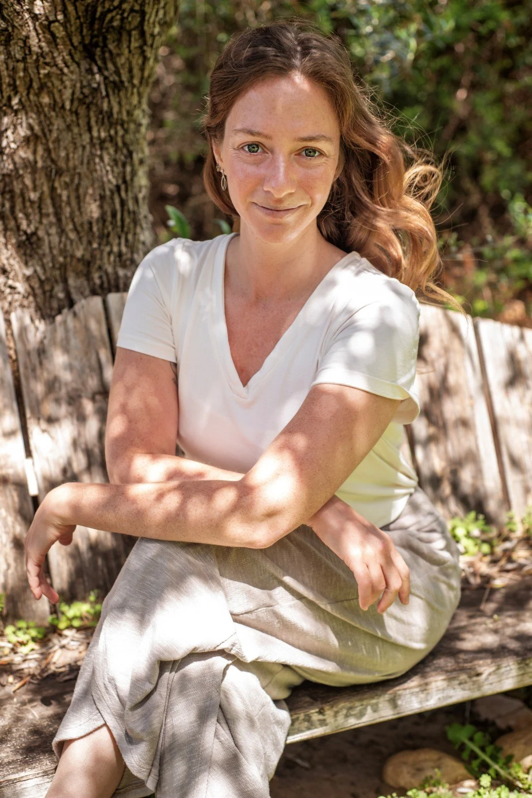 Photo of Rena sitting on a bench with a tree in the background. She is wearing a white t-shirt and smirking at the camera, staring at the camera, hair blowing in the wind