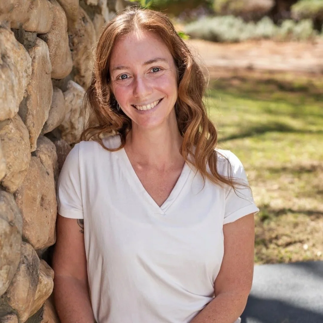 Portrait of Rena wearing a white v-neck tshirt leaning agaist a rock wall with grass in the background, smiling and looking at the camera.
