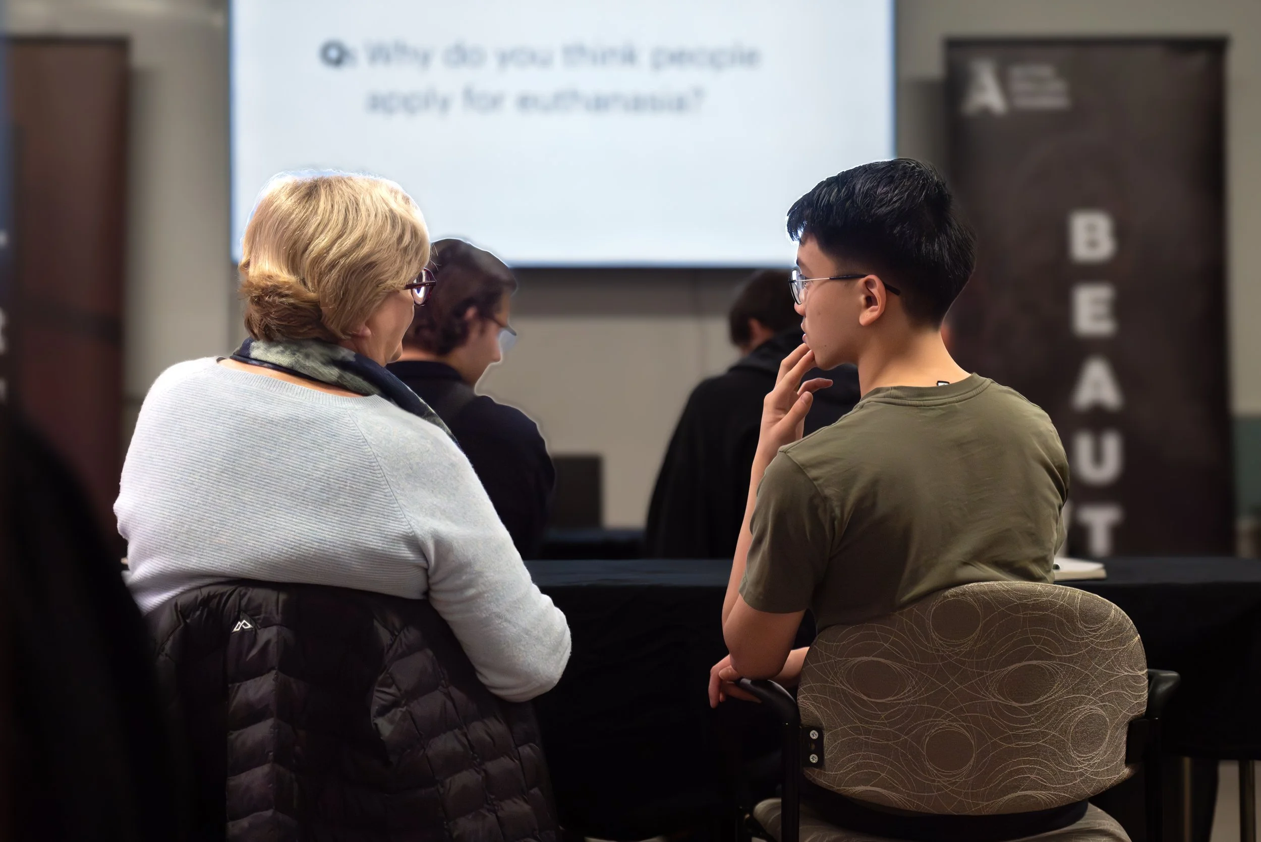 A classroom setting with students seated at desks, one of whom is a young man with glasses, and an older woman with blonde hair listening attentively, with a presentation slide and a 'BEAUX' banner in the background.