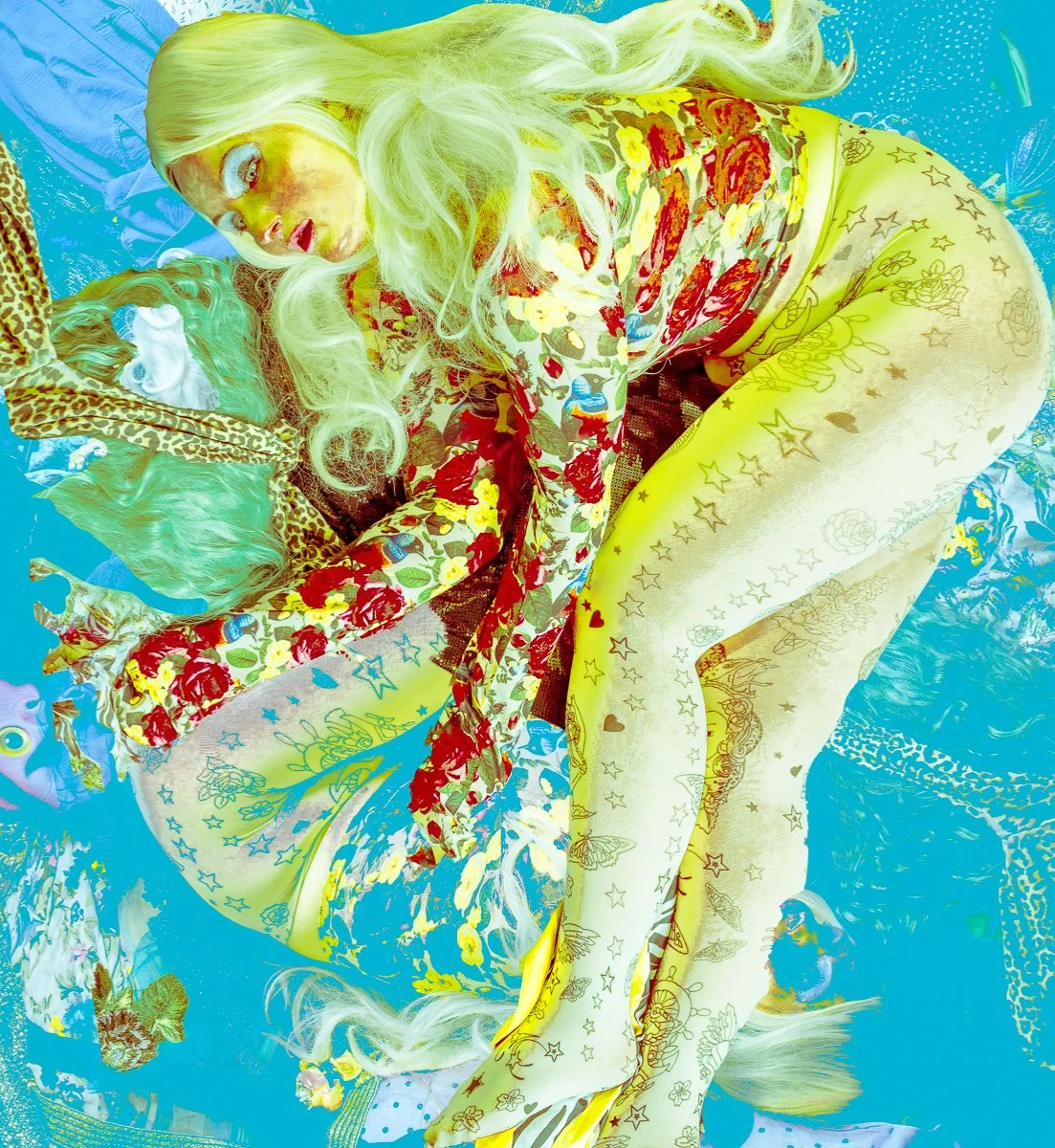 A woman with long blonde hair lying on a colorful, patterned fabric and surrounded by various vibrant, patterned textiles.