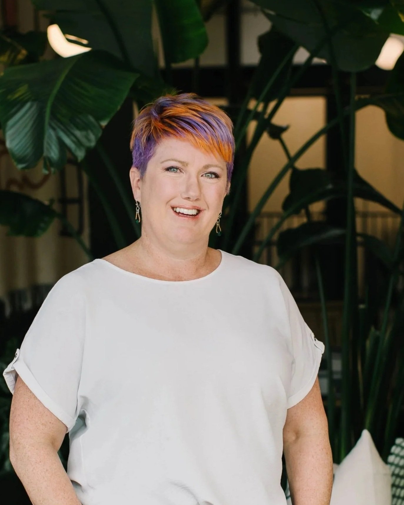 A woman with short, multi-colored hair (purple and orange) smiling at the camera, wearing a white blouse, standing indoors with large green tropical plants in the background.
