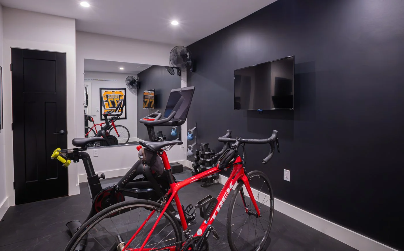 Indoor fitness room with a red racing bicycle, stationary exercise bike, treadmill, wall-mounted television, black walls, mirror, and wall-mounted fans.