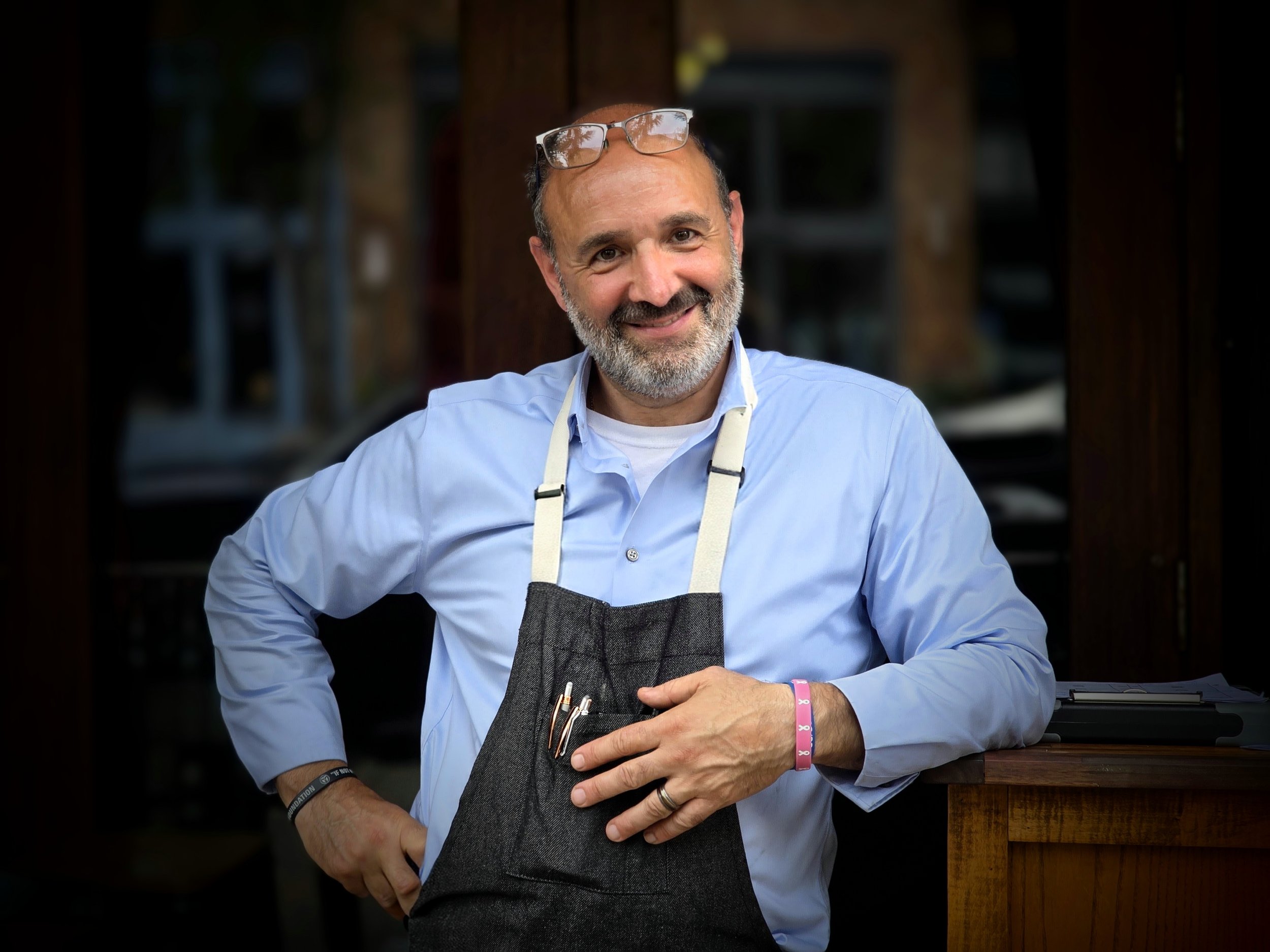 A smiling man with a beard and glasses has them resting on his head while wearing a light blue shirt, an apron with pens, and a pink wristband, standing at a table outdoors.