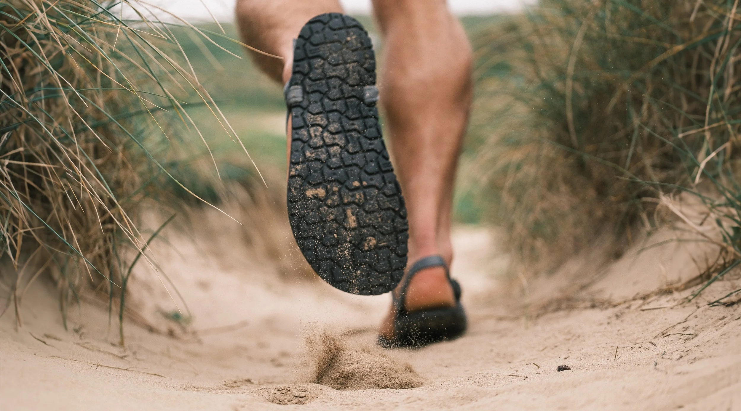 Close-up of a person running on a sandy trail surrounded by tall grass, showing the sole of the shoe kicking up sand.