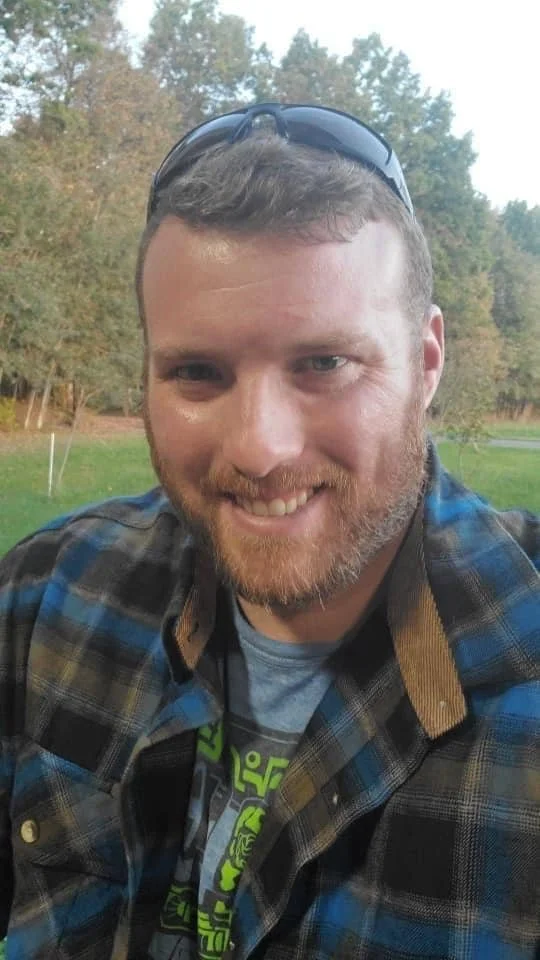 Man outdoors wearing sunglasses on his head, smiling, with trees and a grassy area in the background.