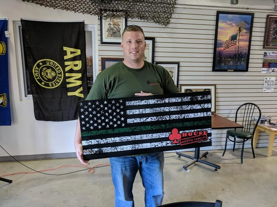A man holding a wooden American flag with a green stripe, featuring a red clover and a 'House Brewing Co.' logo, standing inside a room with military and patriotic decor.
