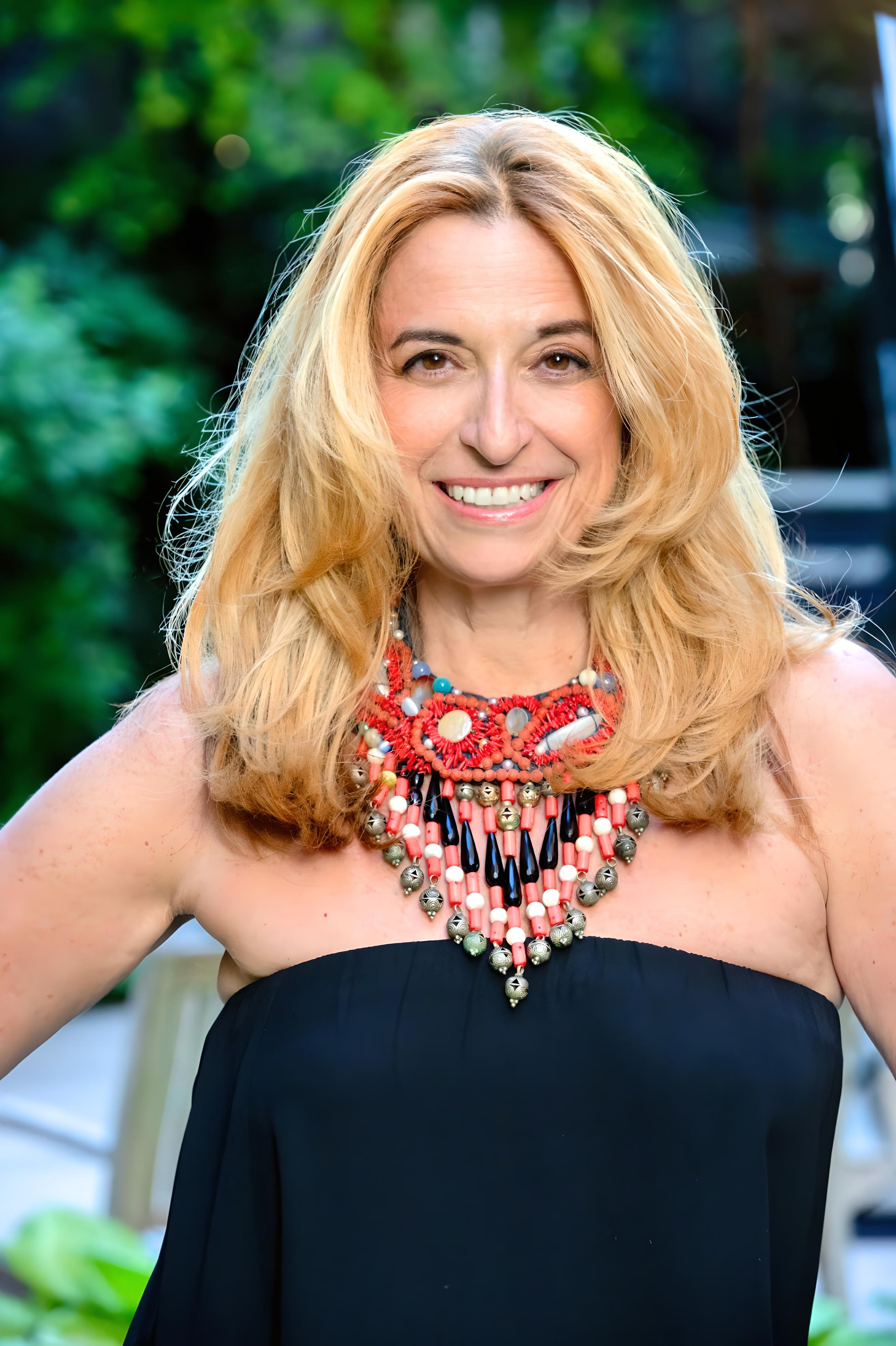 Portrait of Nathalie Botros with blonde hair, standing outside against a green background, dressed in a strapless black outfit and a vibrant red and gold statement necklace.