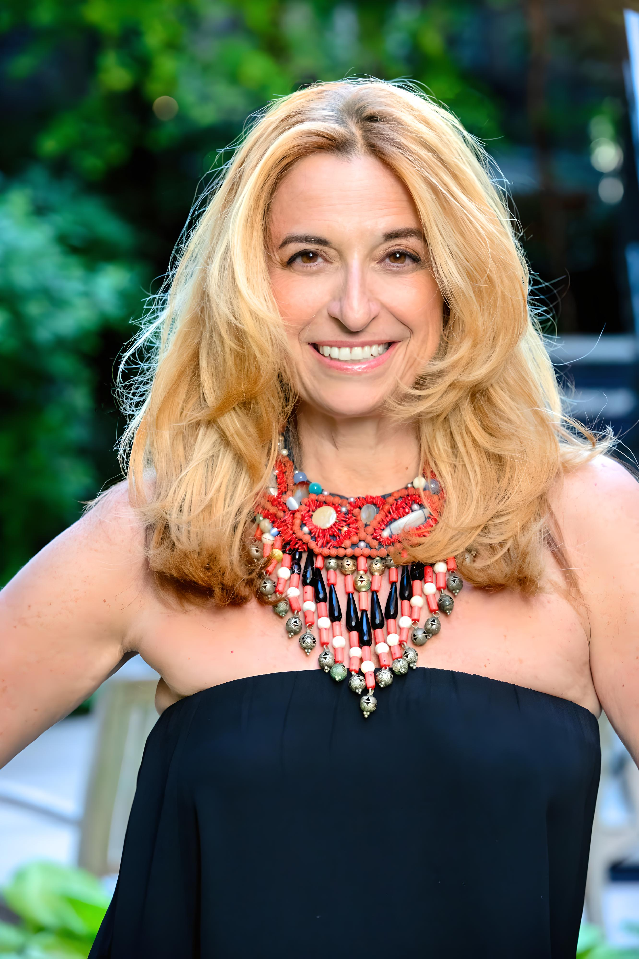 Portrait of Nathalie Botros with blonde hair, photographed outside, dressed in a strapless black top and vibrant red, black, and metallic beaded necklace.