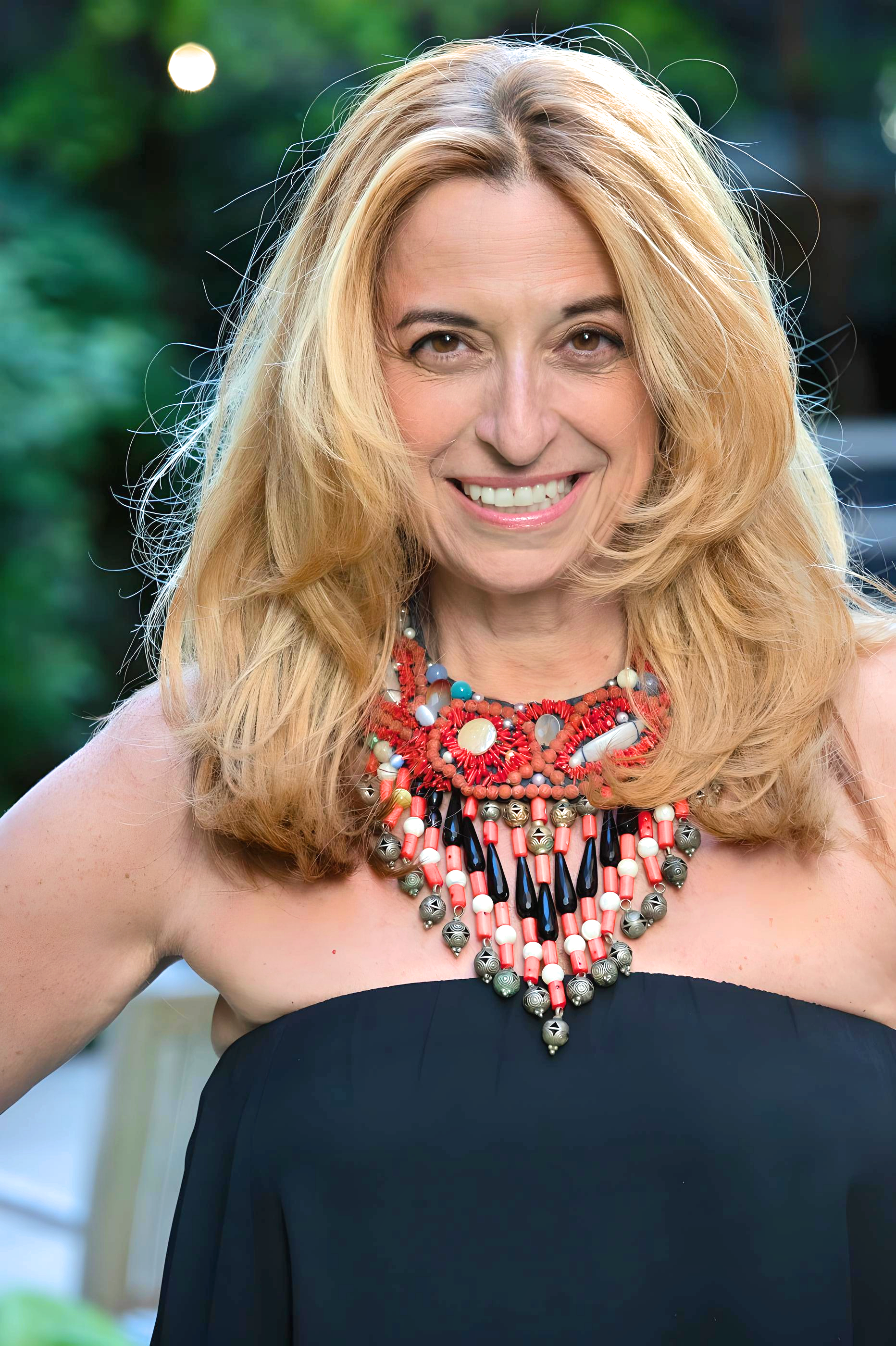 Nathalie Botros smiling outdoors, wearing a strapless black top and a colorful statement necklace, photographed against a green background.