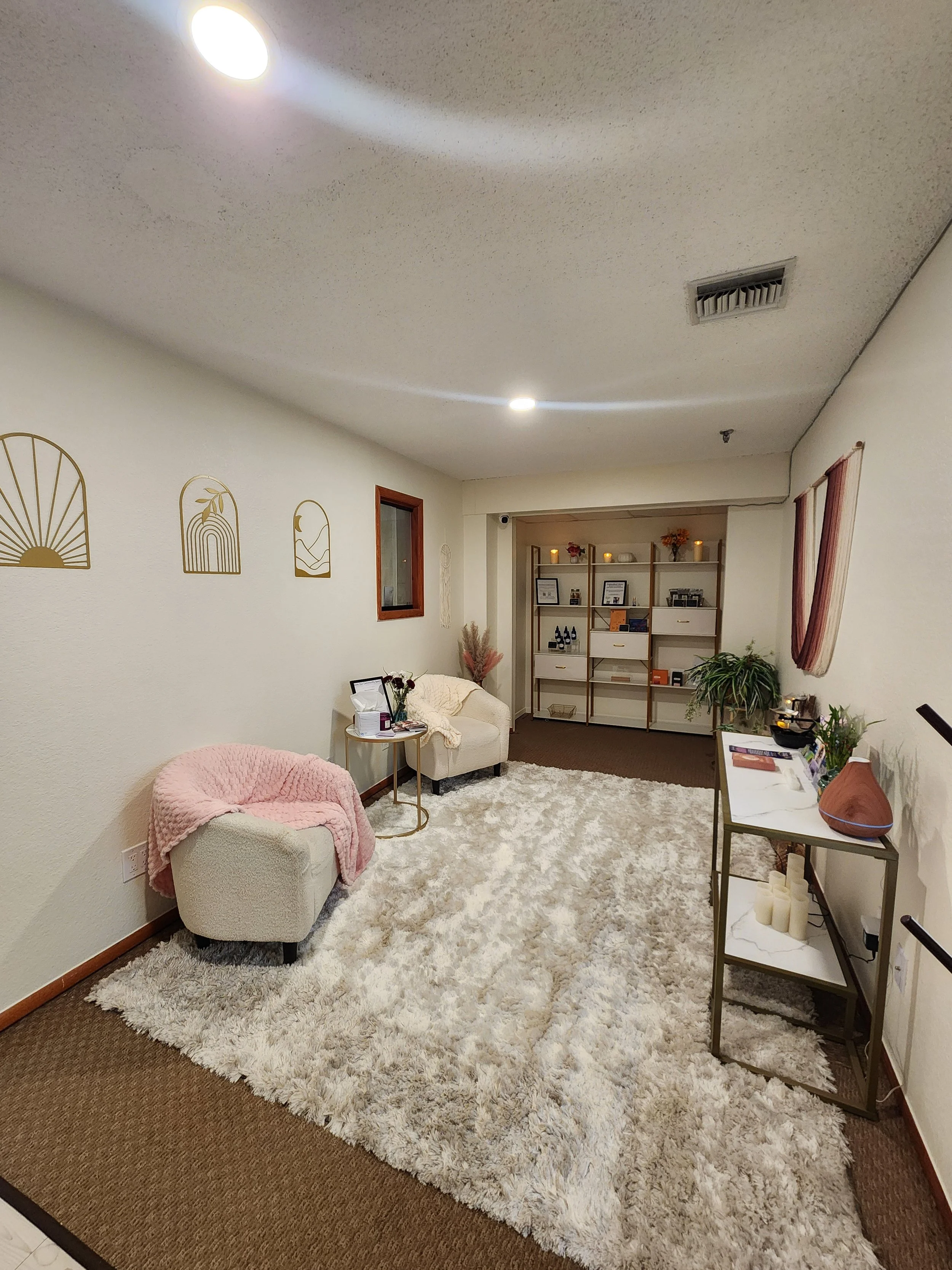 A cozy living room with white walls, a fluffy beige rug, and a pink armchair. A small white armchair has a pink blanket draped over it. Decor on the walls includes gold wall art. A wooden shelving unit holds framed pictures, candles, and decorative i