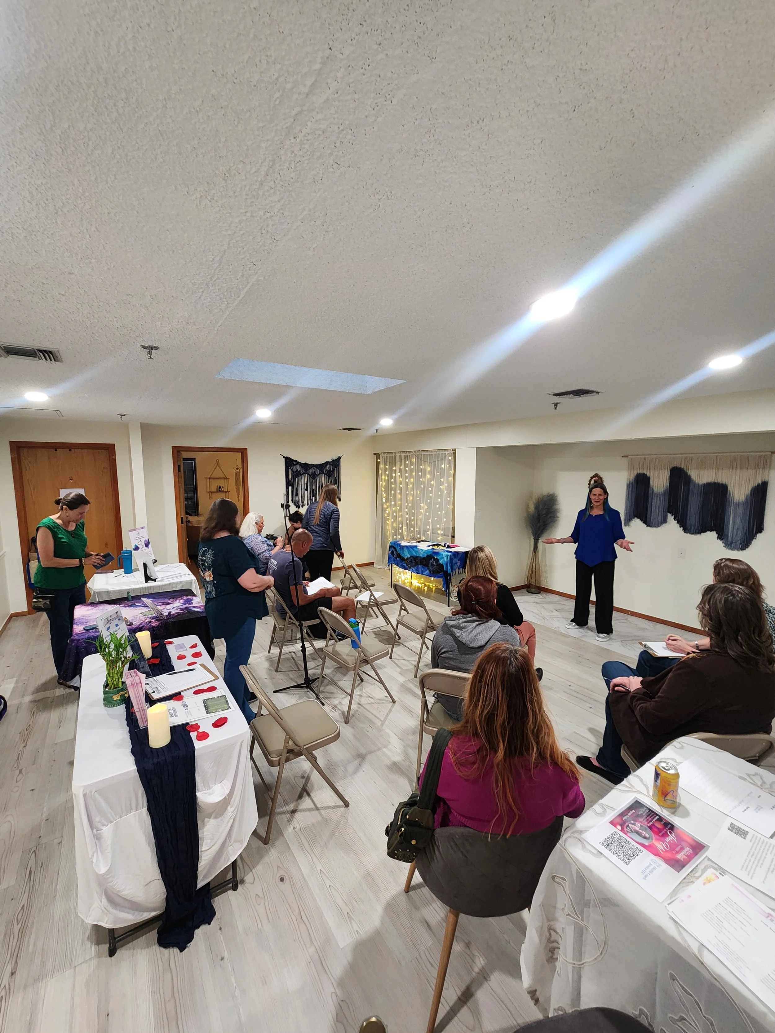 An indoor gathering with people seated and standing around tables, a woman standing at the front speaking to the group, tables with informational materials, and festive decorations on the wall.