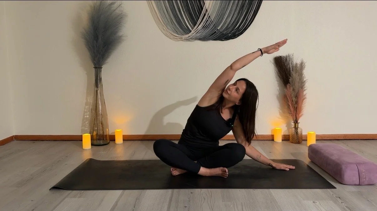 Woman practicing yoga in a peaceful room decorated with candles, vases with pampas grass, and wall art, sitting in a seated twist pose on a black yoga mat.