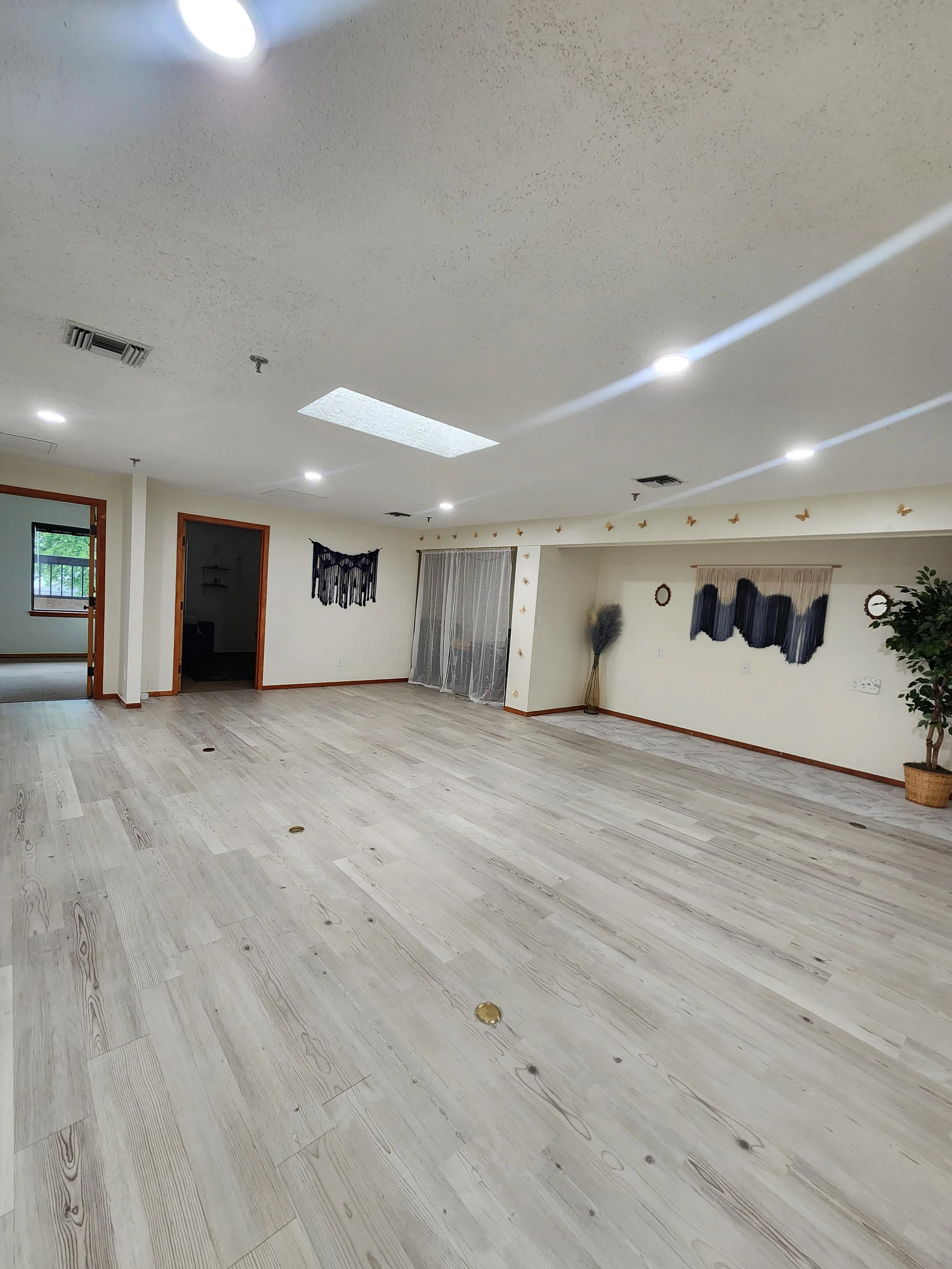 Empty room with light-colored wood flooring, white walls, doorways, window, and decorative wall hangings, illuminated by ceiling lights.