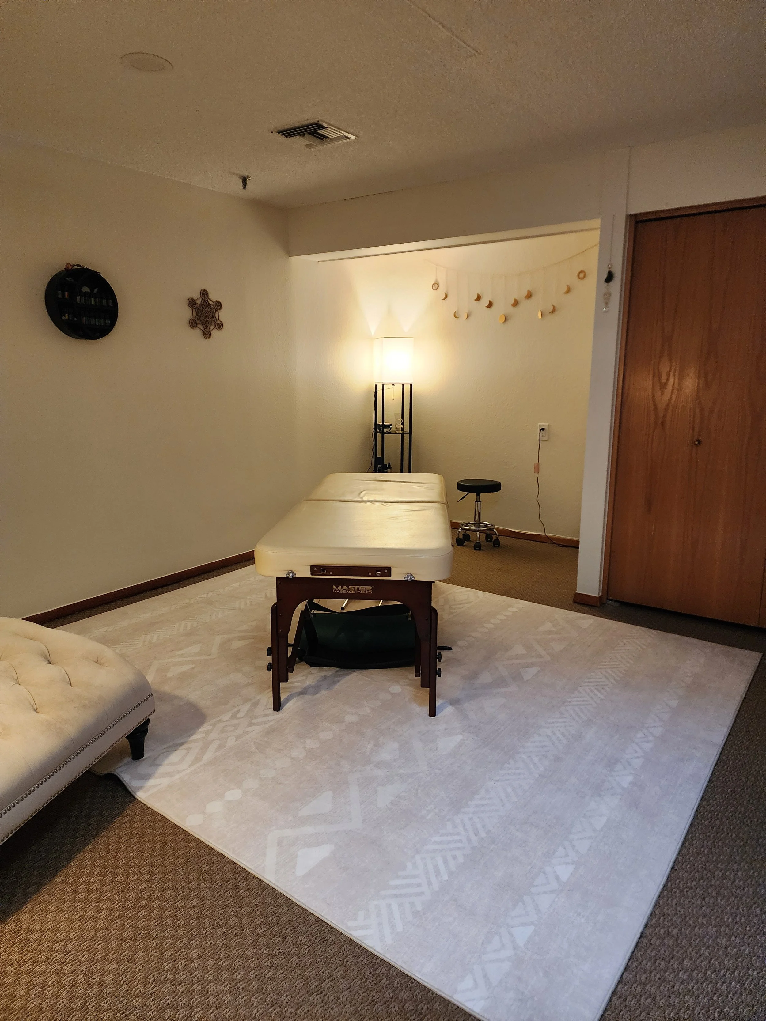 A room with a textured ceiling, beige carpet, and white walls. There are wall decorations, a colorful patterned rug, and furniture including a white tufted couch, a cream bed, a black chair, and a tall black shelving unit with a lamp behind it. There are string lights and small hanging decorations in the corner.