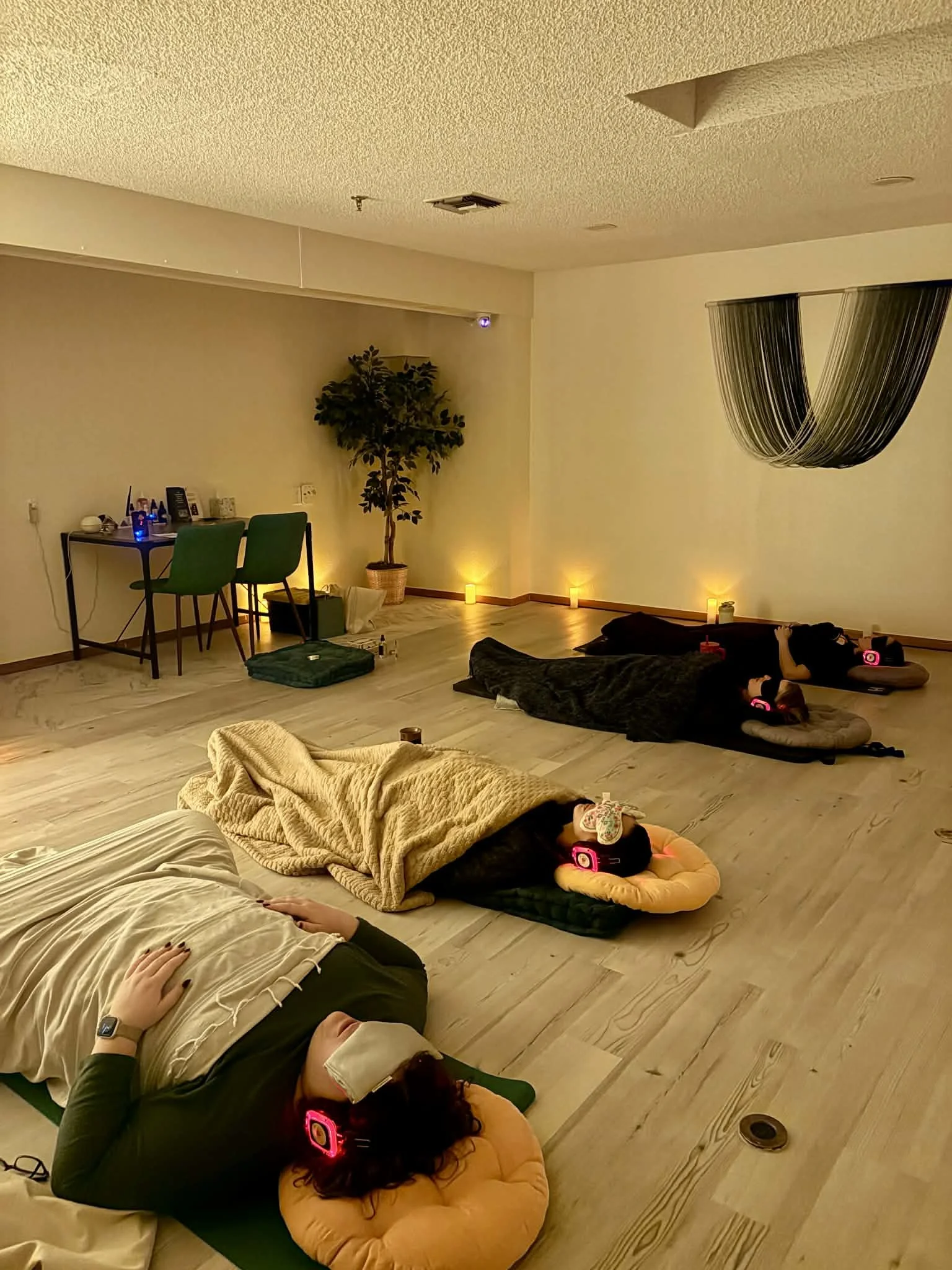 People are lying down on yoga mats or cushions, listening to music with headphones, in a dimly-lit room with candles and a decorative hanging on the wall.