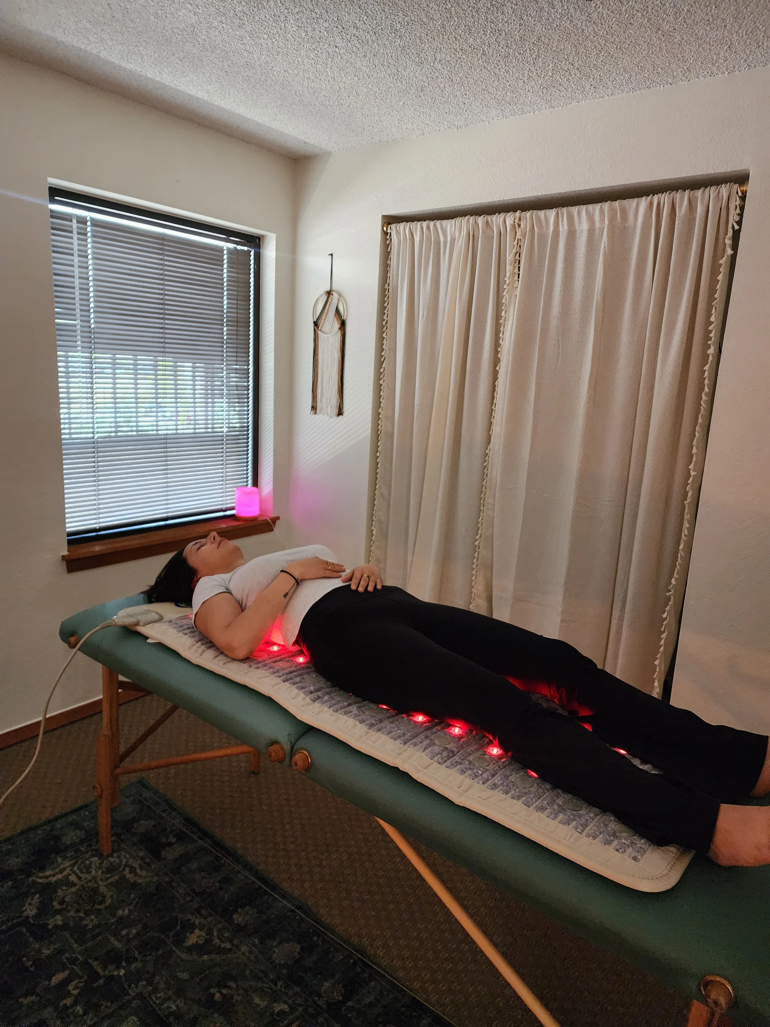 Woman lying on a massage table with red light therapy pads underneath her, in a room with a window and beige curtains.