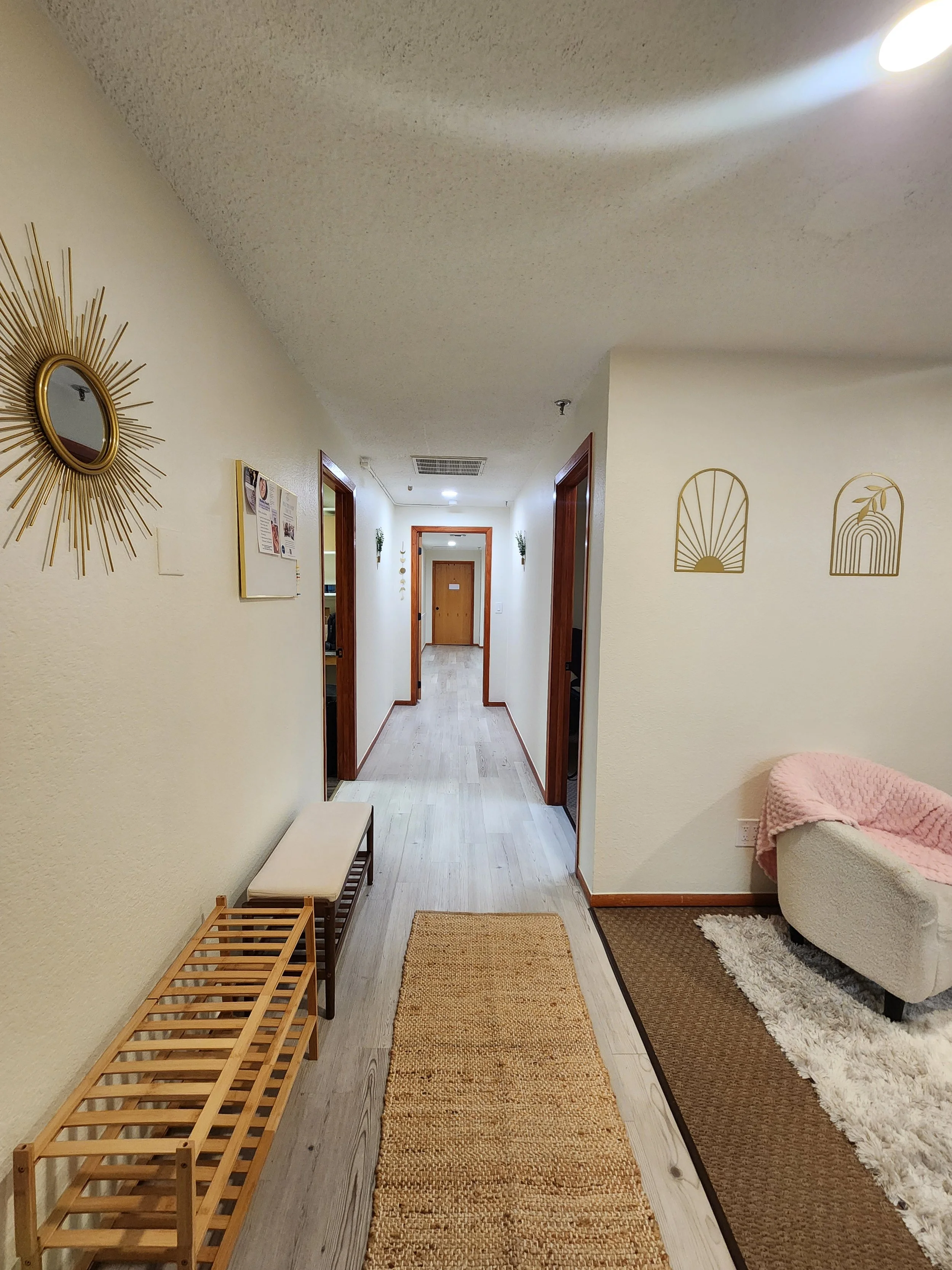 A hallway with light beige walls, a wooden door at the end, and two open doors on either side. Decor includes gold wall art, a mirror with a sunburst frame, a pink blanket on a white couch, a woven rug, and a bench with a slatted wooden design.