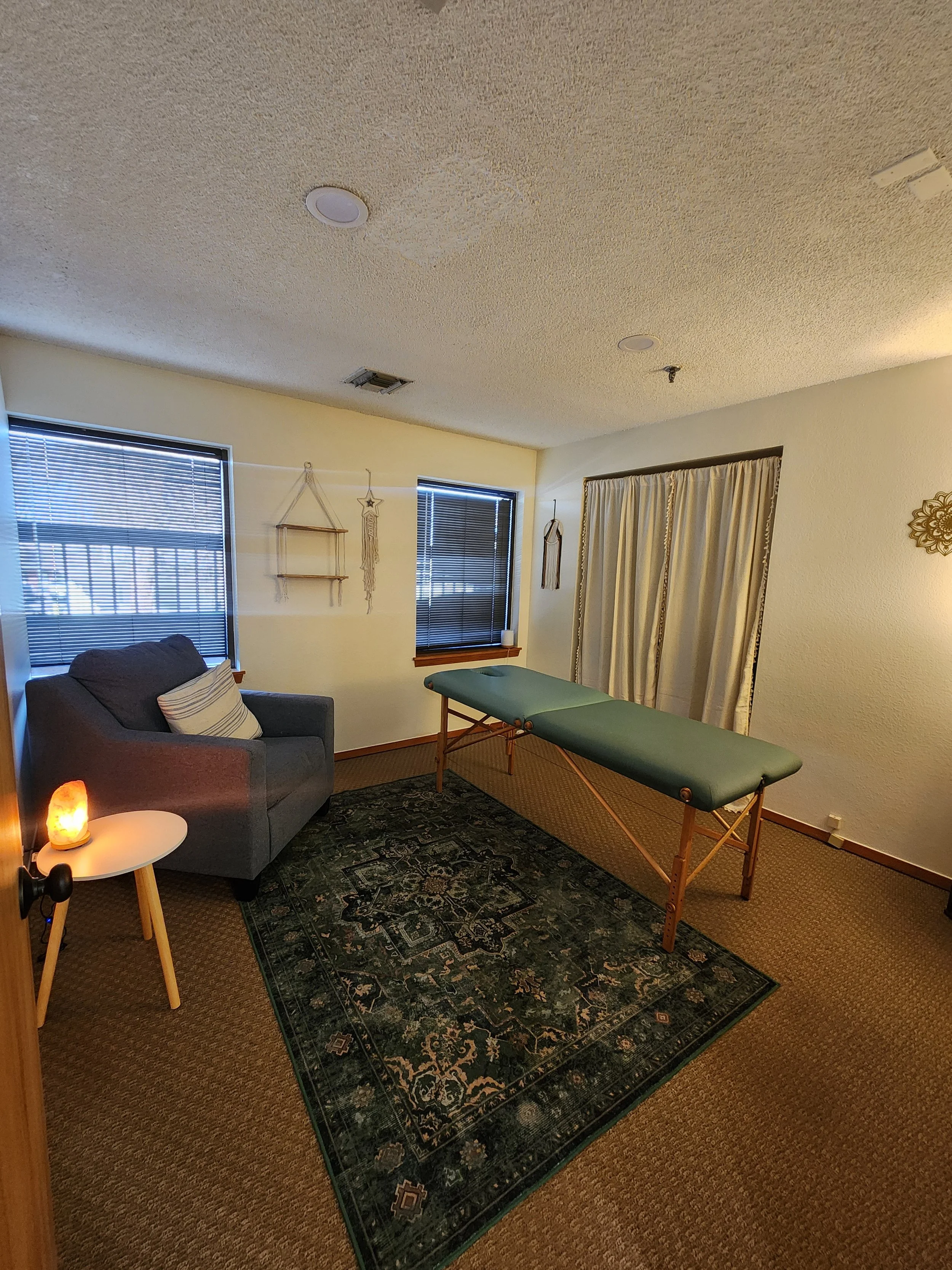 A massage room with a massage table, a gray armchair, a small side table with a salt lamp, two windows with blinds, wall decorations, and a curtain covering an entrance.