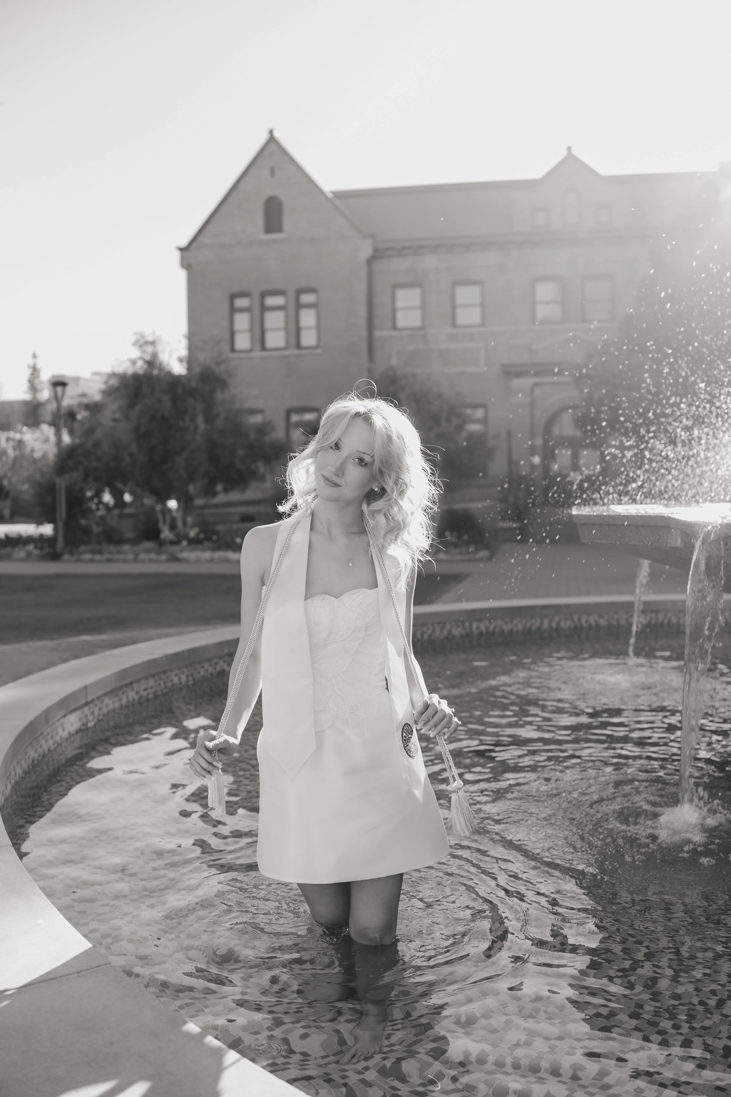 Female ASu graduate taking a celebratory photo in the fountain at Old Main on campus. Wearing a white graduation dress & graduation stole. Black and white image.