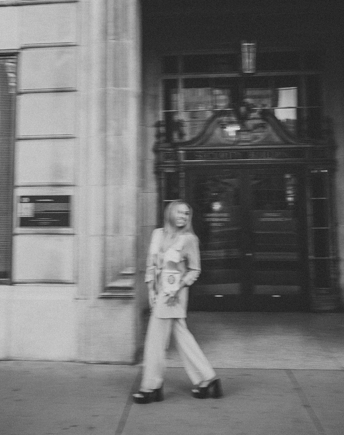 A vibey aesthetic image (slightly blurred) in downtown phoenix, of an ASU graduate. She is walking to the left but looking right. In a very cool girl-chic pant suite and her stole. The building offers cool old-school architecture and a stand-out door