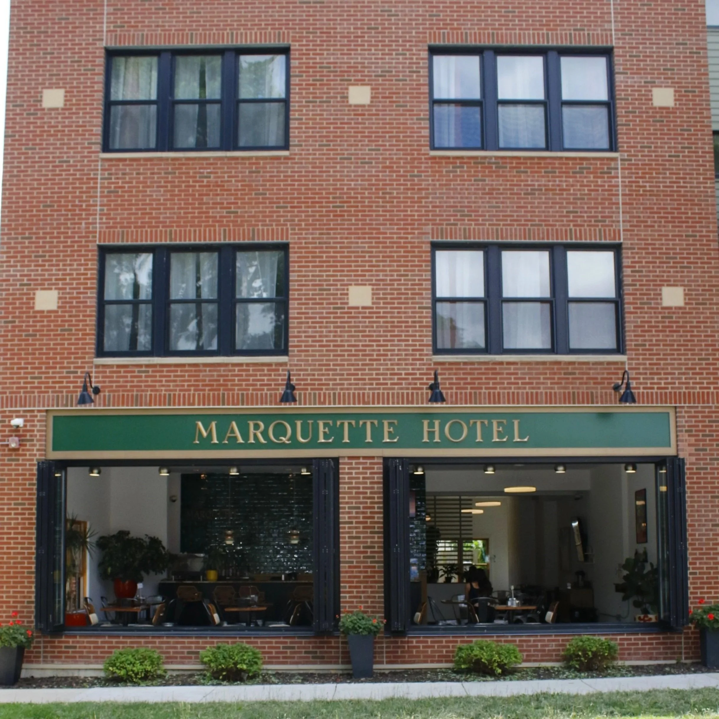 The exterior of the Marquette Hotel, a brick building with large windows and an open ground-floor restaurant or lounge area with outdoor seating.