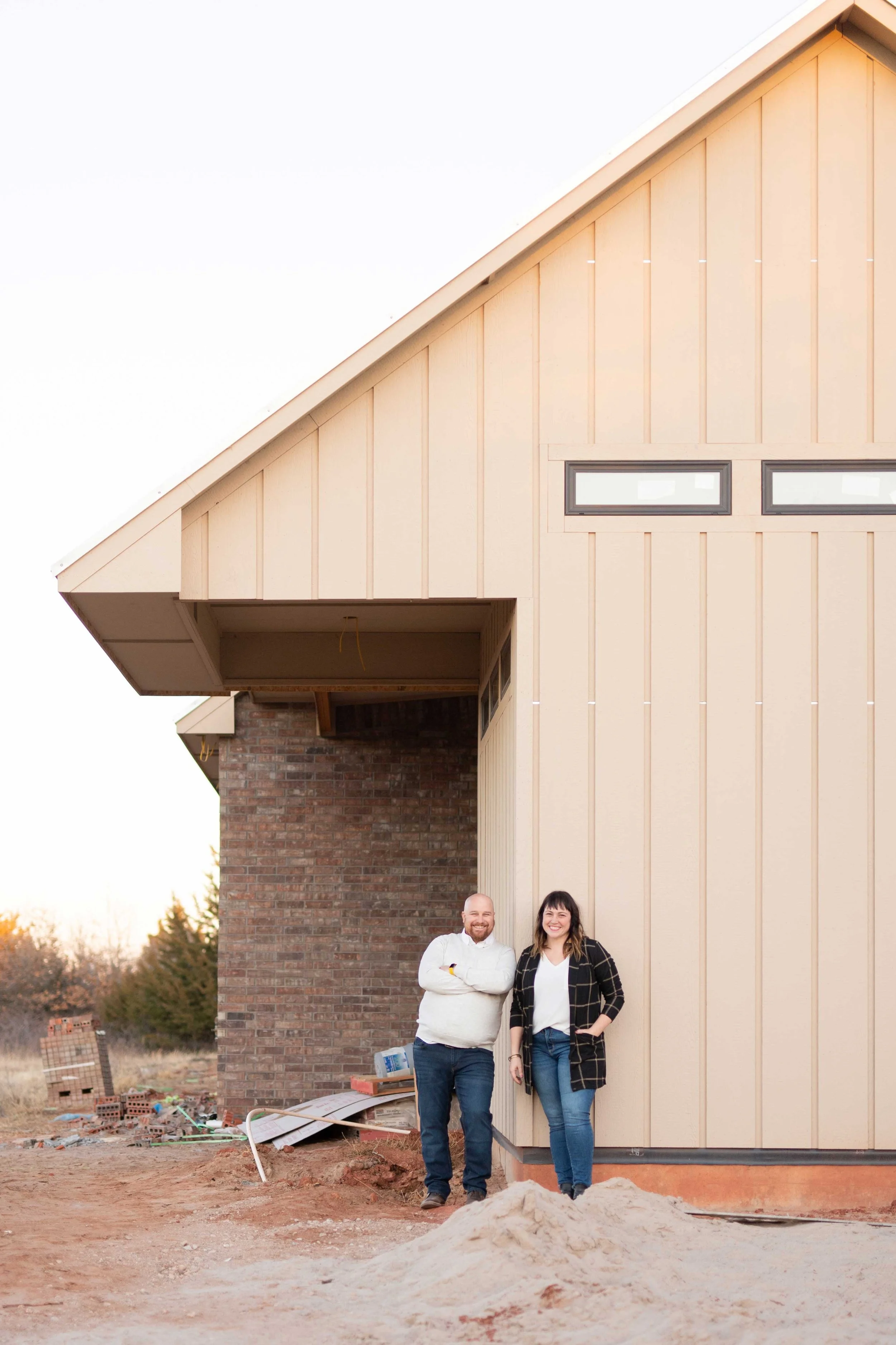 Two people standing in front of a house under construction, smiling at the camera. The house has horizontal beige siding, a brick section, and several small windows. The ground is dirt with some construction materials scattered around.