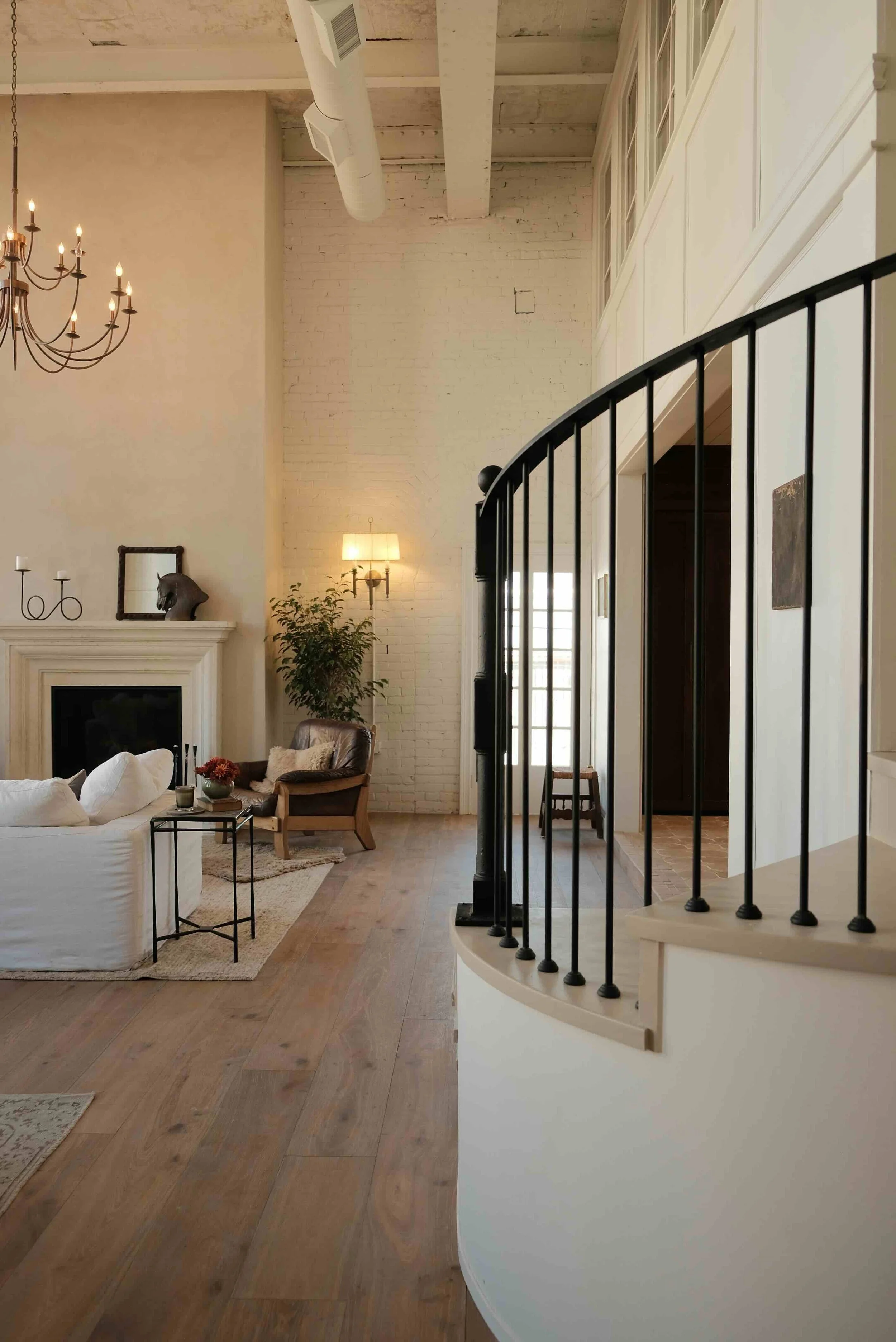 A spacious living room with a high ceiling, exposed white brick wall, and wooden flooring. It features a black metal staircase railing, a white sofa, a chair with a throw pillow, a fireplace, and decorative items such as a chandelier, candle holders, a horse sculpture, and a potted plant. There's also a wall-mounted lamp and a window allowing natural light.