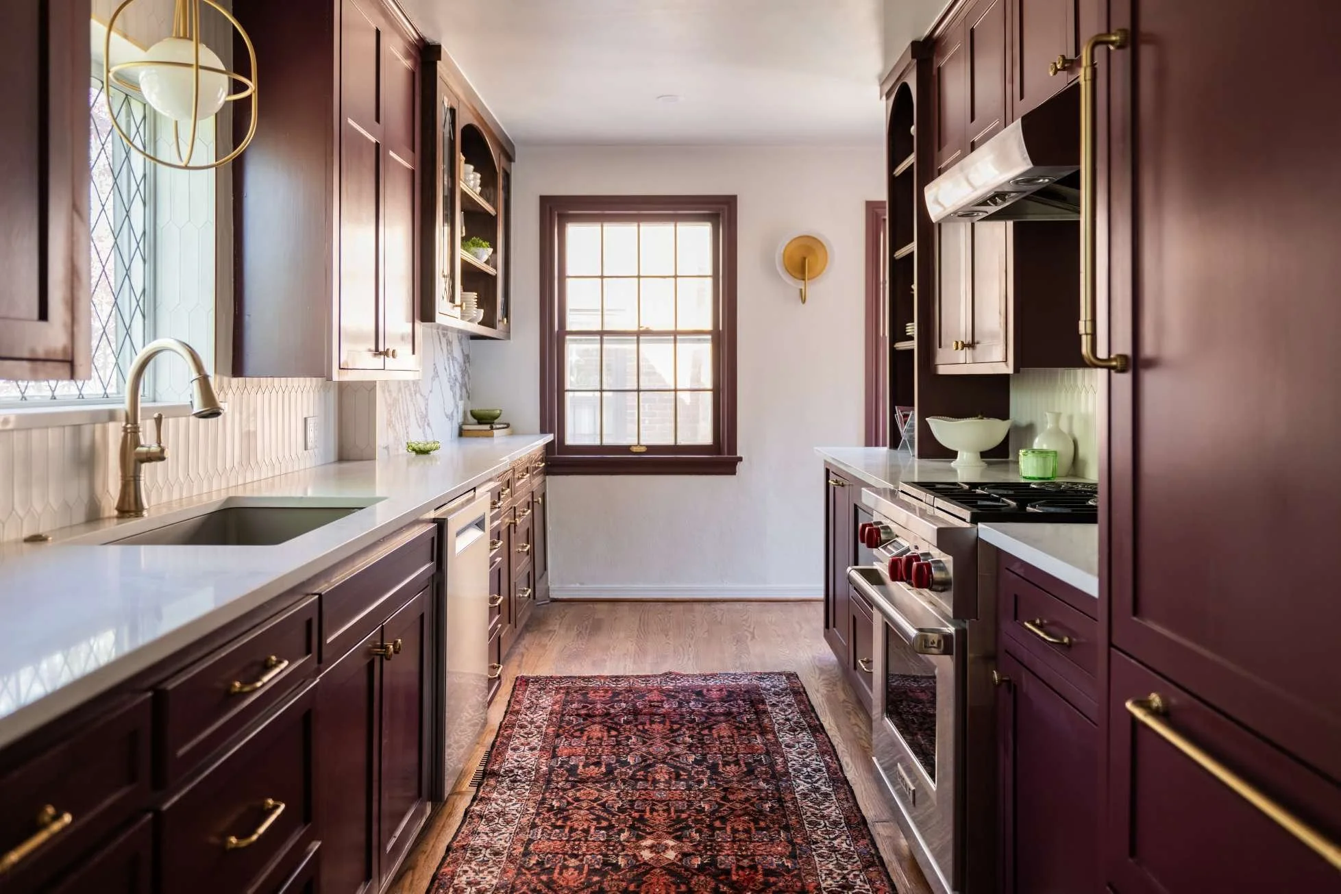 A kitchen with dark purple cabinets, white countertops, and a patterned rug. It features a window with sunlight, a modern gold and glass light fixture, and a backsplash with white tiles. There’s a sink under the window and a stove on the right side, with open shelves and decorative bowls on the counters.