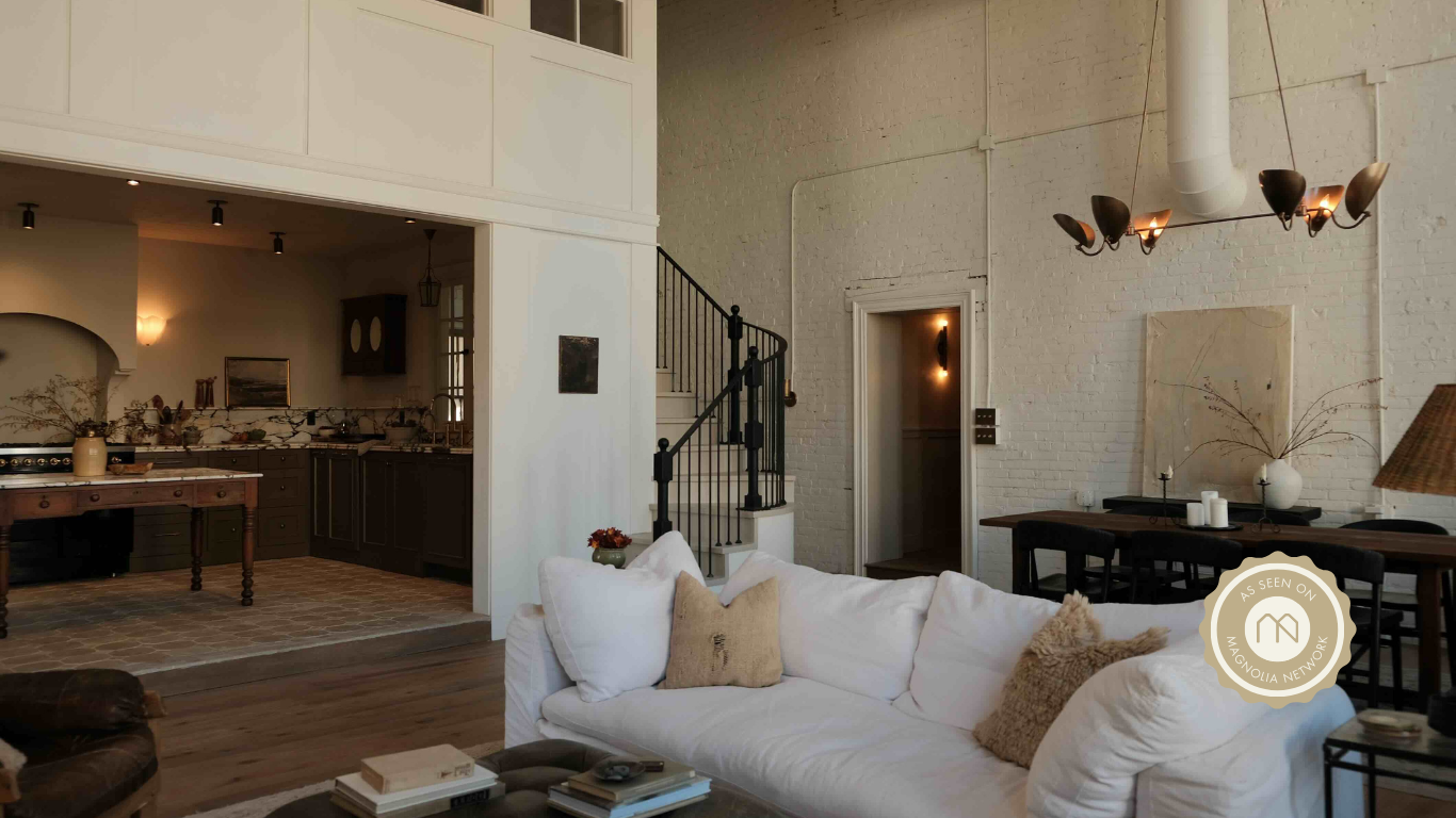 Living room area with white brick wall, black dining table and chairs, white sofa with beige pillows, staircase with black railing, and a doorway leading to a kitchen with dark cabinets and a marble countertop.