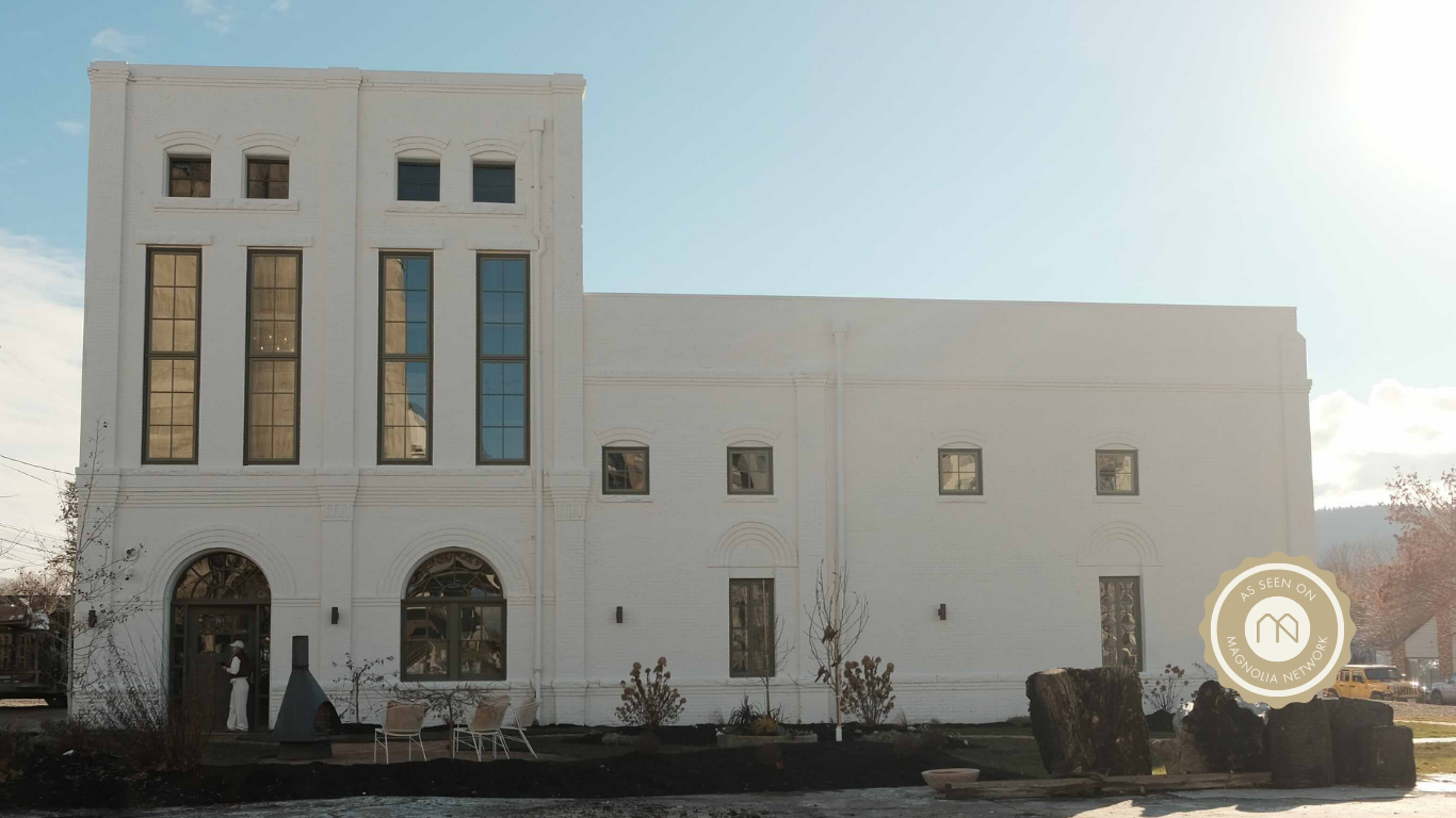 White multi-story building with large and small windows, small trees, and outdoor seating area with rocks and a person near the entrance, under a bright sky.