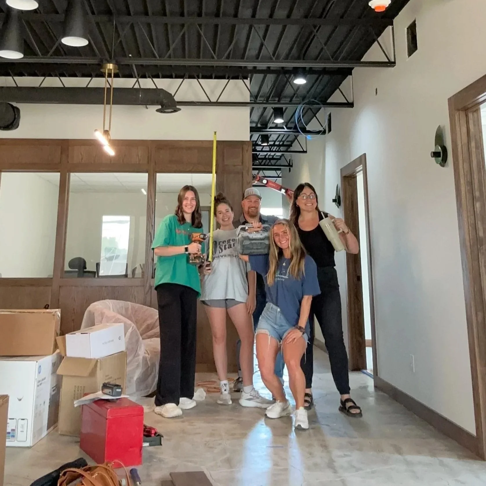 Group of five young adults standing inside a room, smiling, with construction tools and boxes around, indicating a renovation or moving in activity.