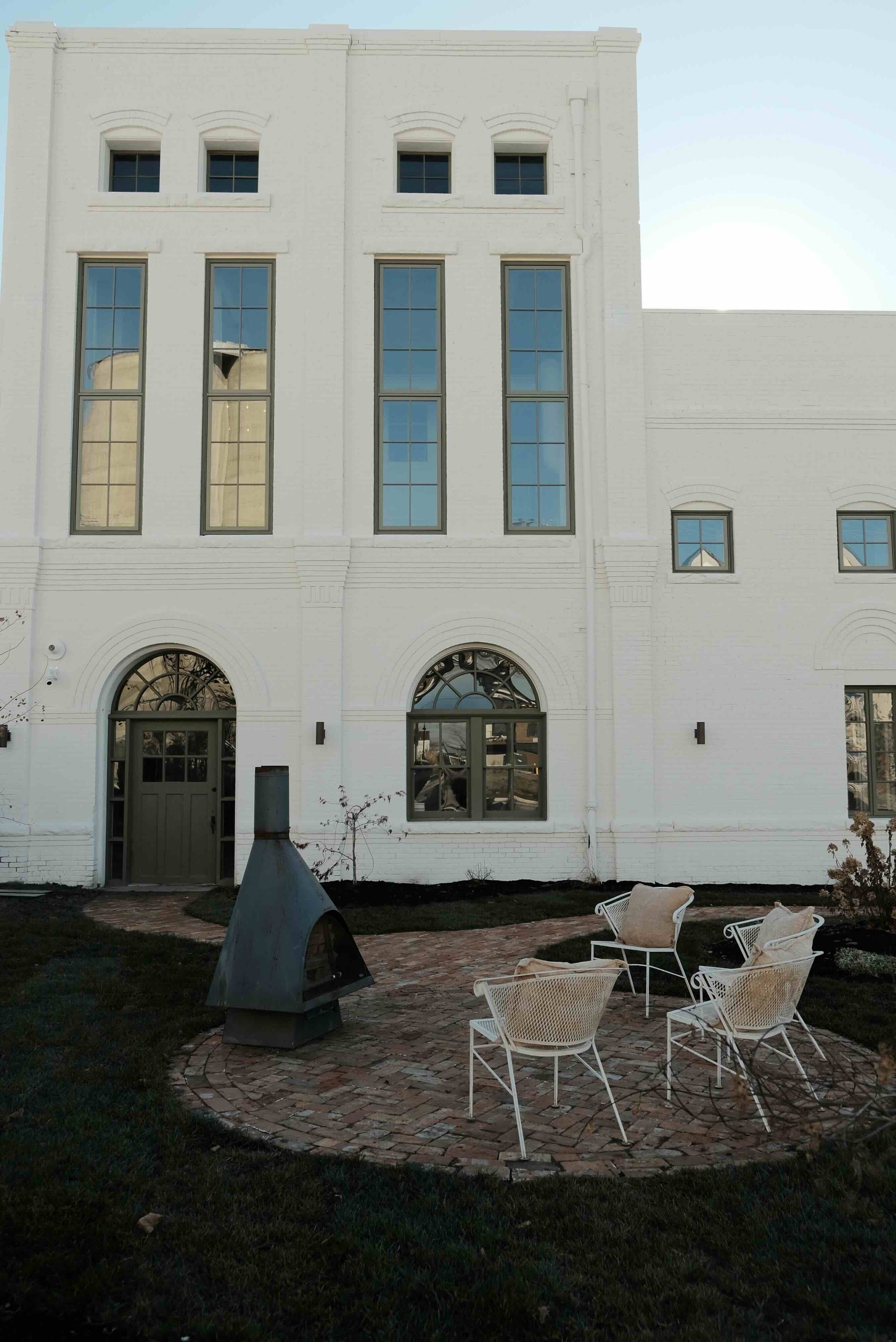 White multi-story building with large and small windows, a brick patio with white chairs and a metal outdoor fireplace in the yard.