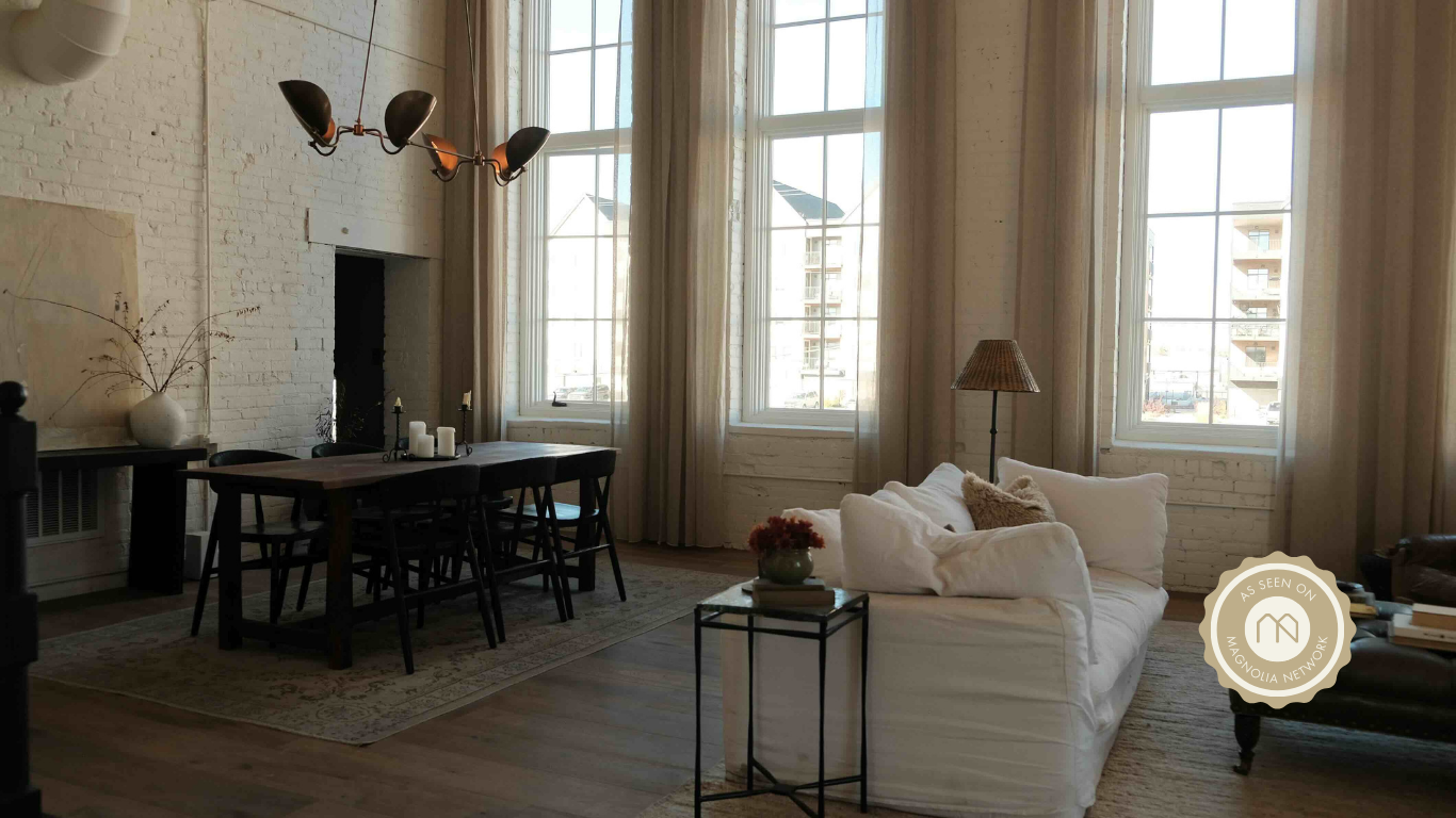 A bright living room with large windows, white brick walls, a white sofa with pillows, a side table with flowers, a dining table with chairs, a chandelier, and a lamp.