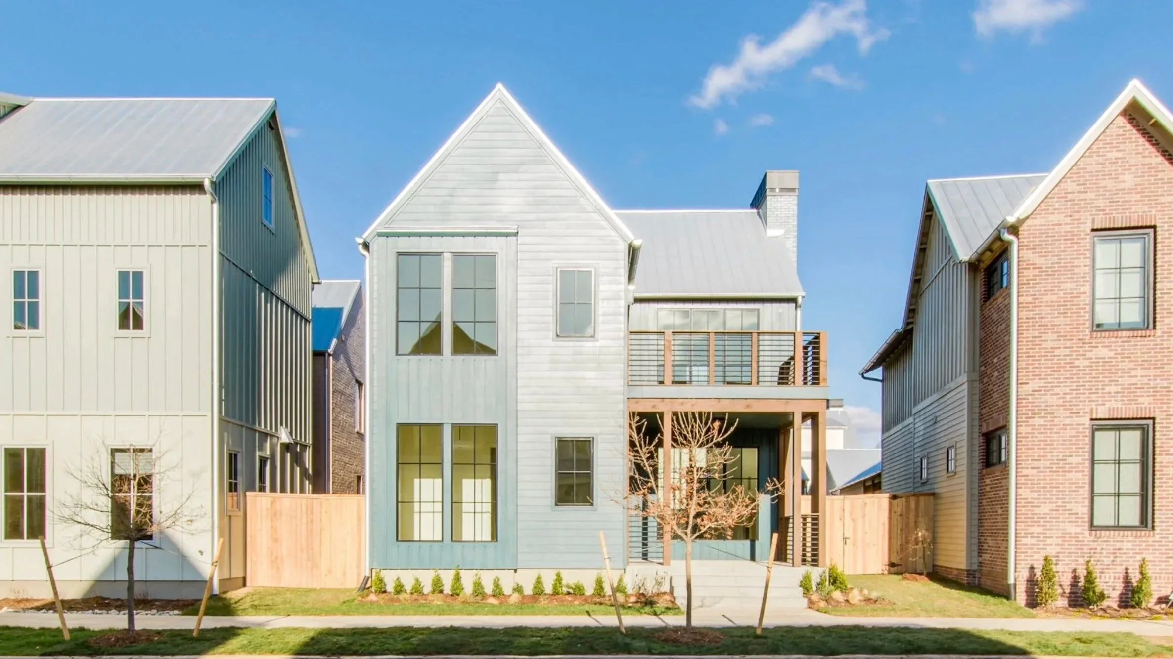 Row of modern houses with different siding and roofing styles, small trees in front, clear blue sky.