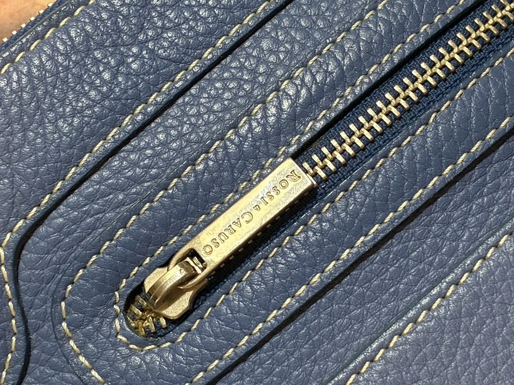 Close-up of brass zipper and engraved Rossi & Caruso logo pull on blue pebbled leather bag.