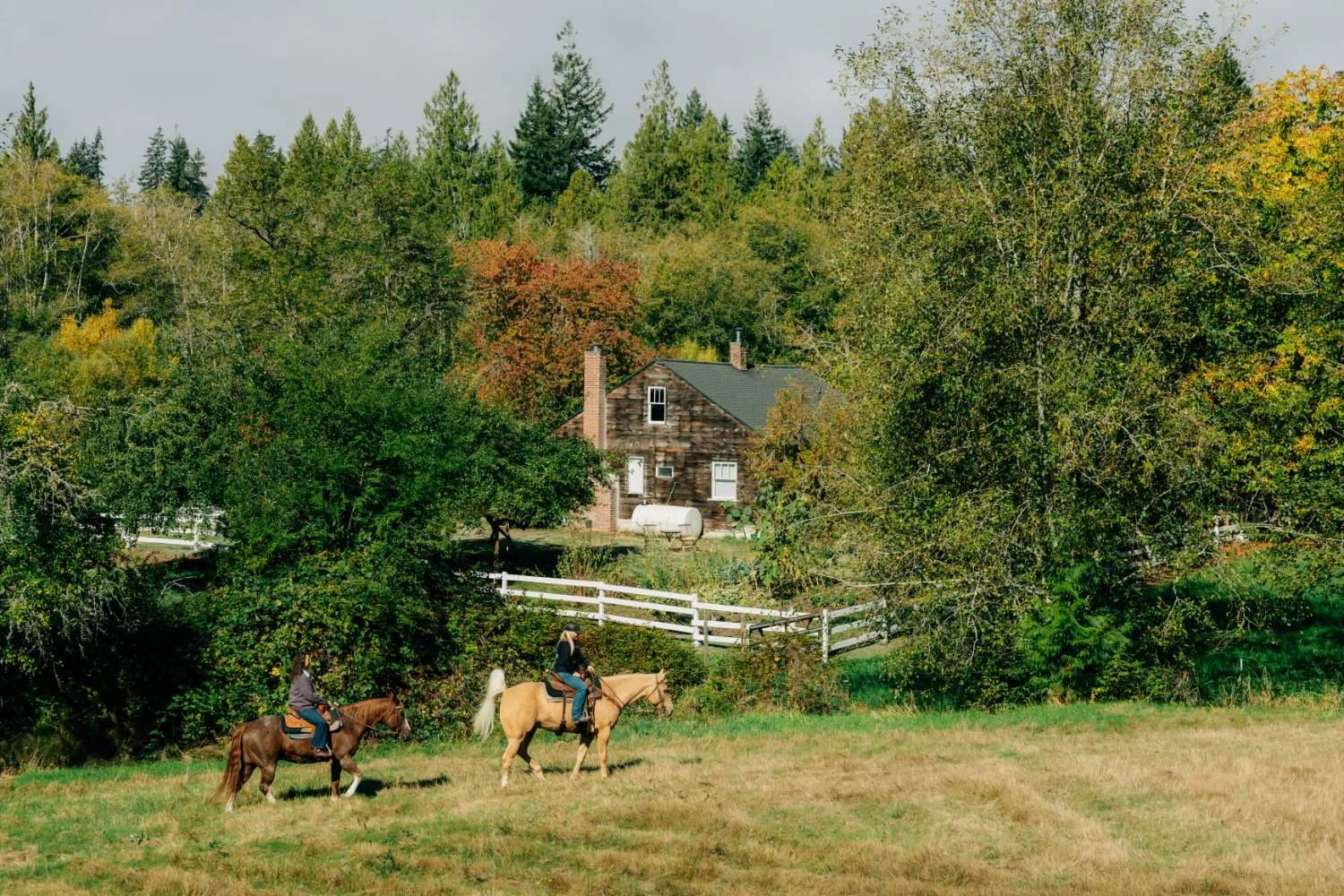 Two riders on horseback with a stunning view of a historic farmhouse in the background