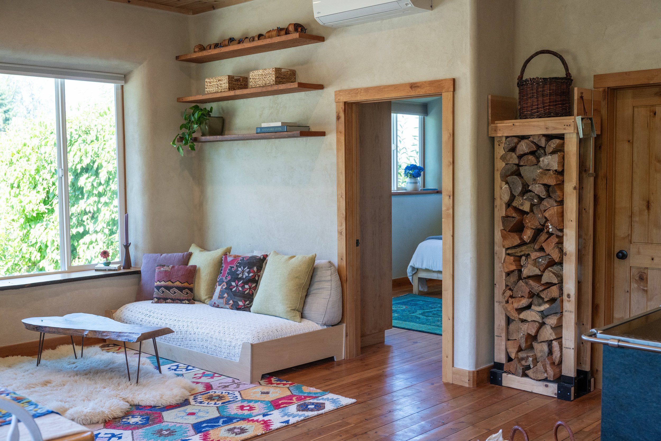 Great room inside the handcrafted straw bale home with clay plaster walls, floating wood shelves, built-in firewood storage, and abundant natural light at Be Easy Farm in Sequim, Washington.