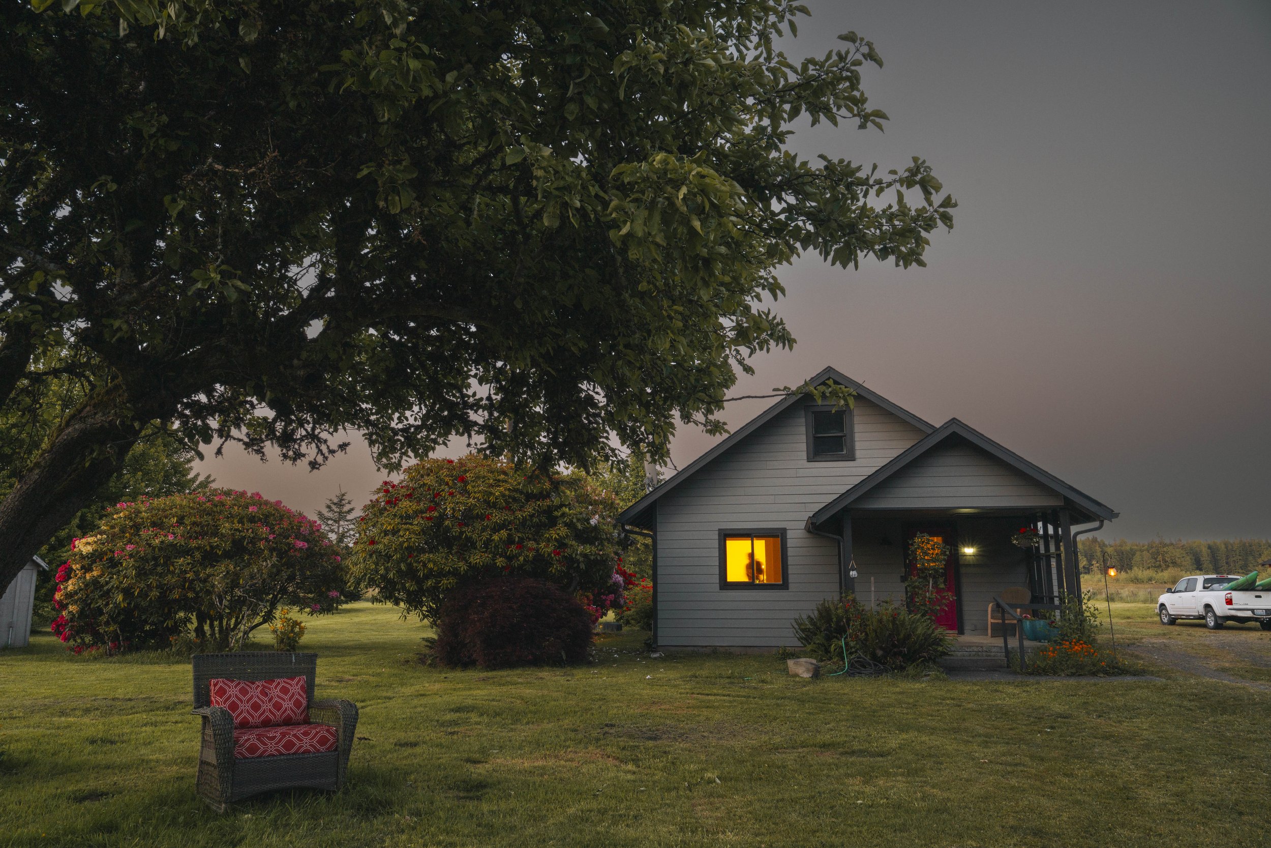 A peaceful summer evening on the farm in Western Washington. a farmhouse in the background, and a garden chair sits under the shade of a large tree