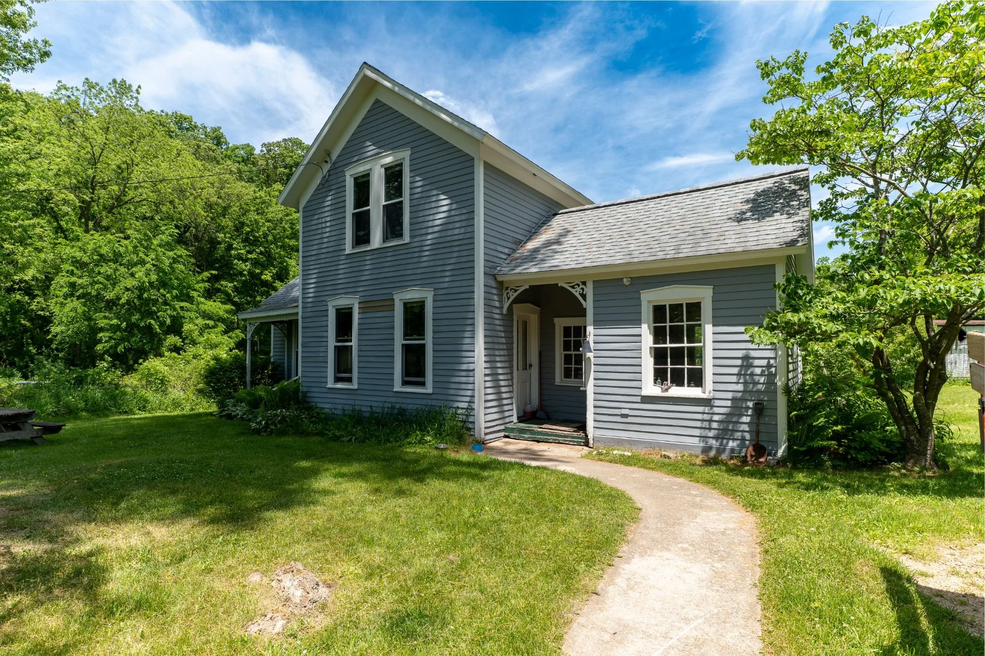 A well-kept farmhouse with tall windows and a cosy front porch. Lush forest in the background.