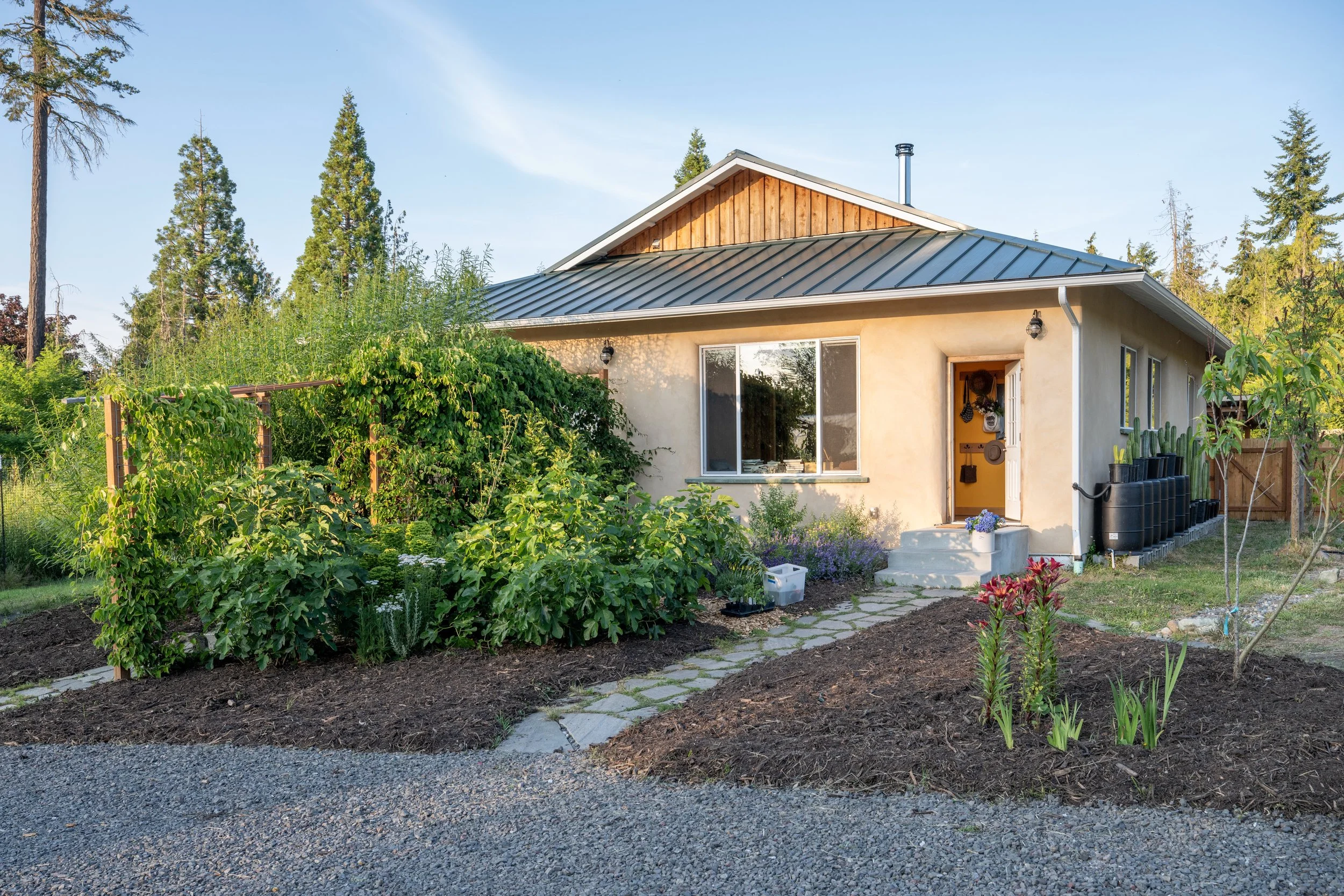 Front exterior of the handcrafted straw bale home at Be Easy Farm, featuring clay plaster walls, a standing seam metal roof, edible landscaping, and mature gardens in Sequim, Washington.
