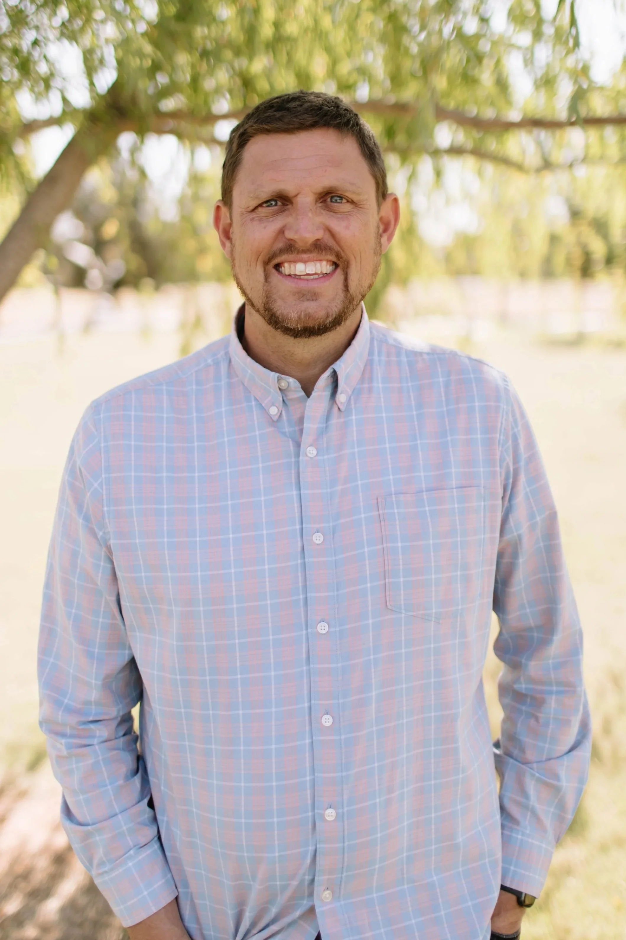 A man with short brown hair and blue eyes, smiling, wearing a blue and white checkered shirt, standing outdoors in front of a tree with green leaves and a grassy background.