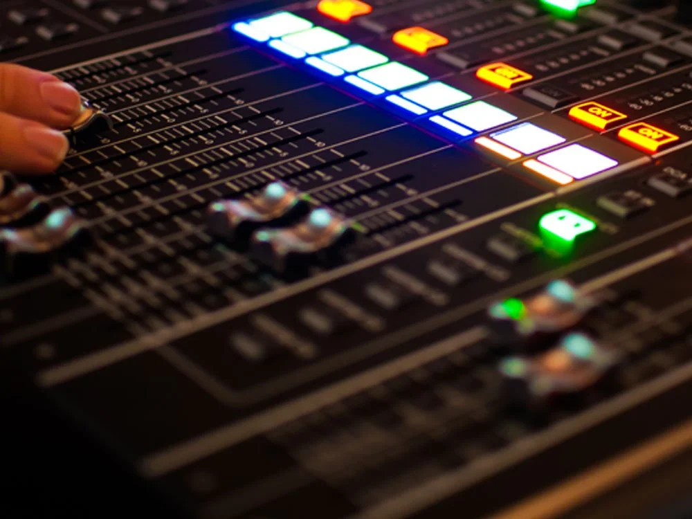 A close-up of a sound mixing console with illuminated faders and controls, including green, blue, and red lights.